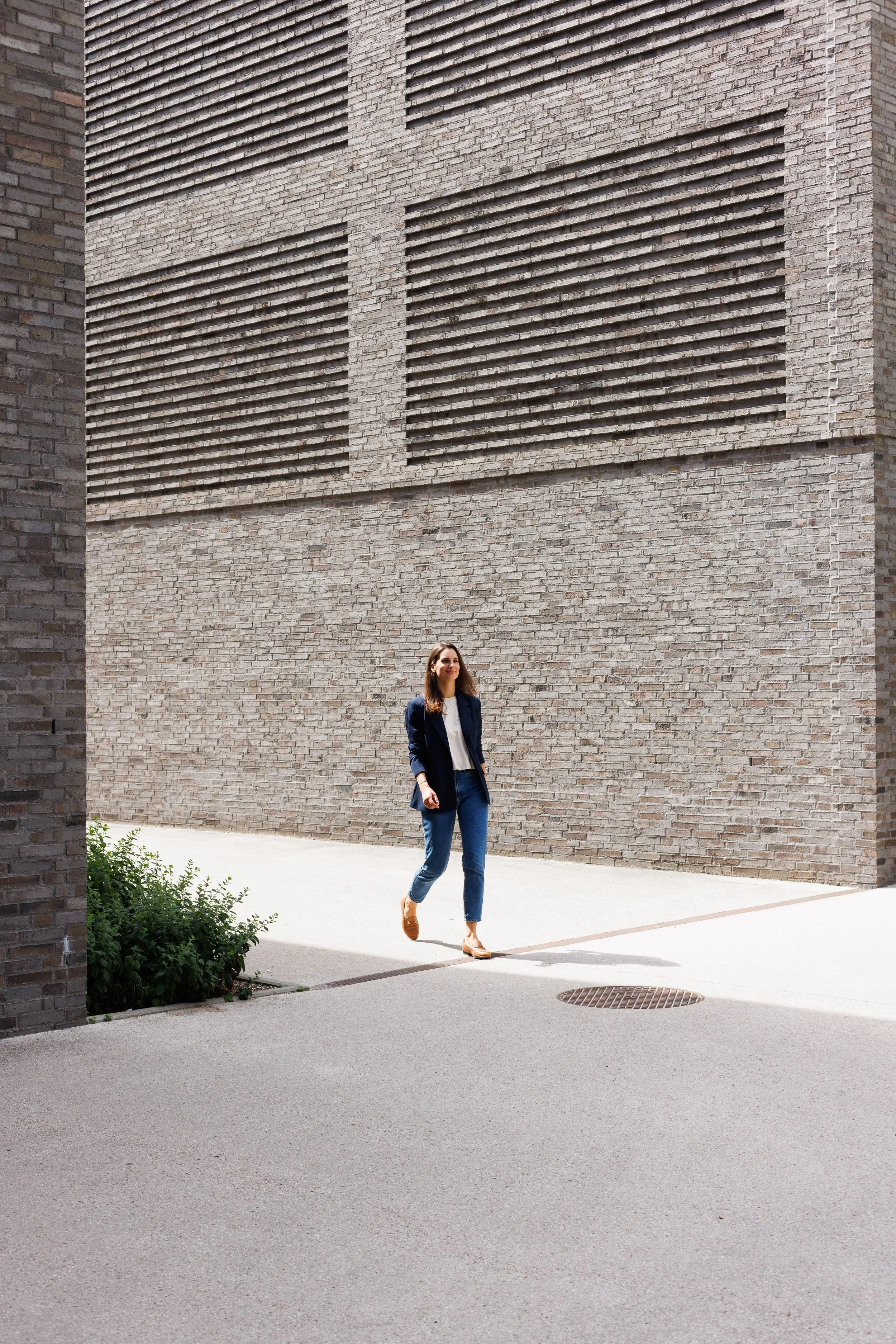 Frau in blauer Blazer, hellblauer Jeans, braunen Schuhen beim Gehen auf einer Straße vor einer großen Backsteinwand mit Lüftungsschlitzen.