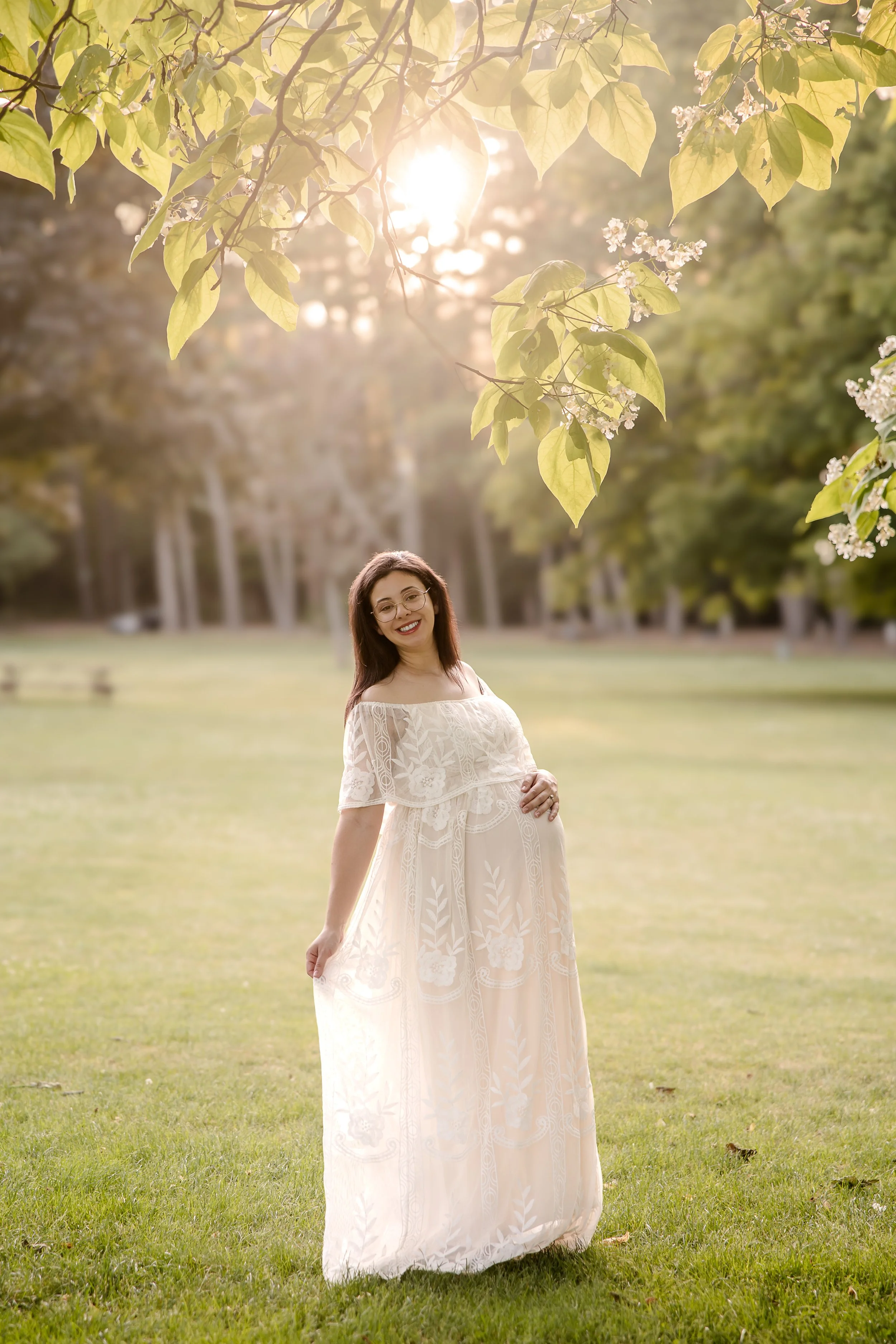 smiling pregnant woman in a white dress standing outside with the sun shining behind her
