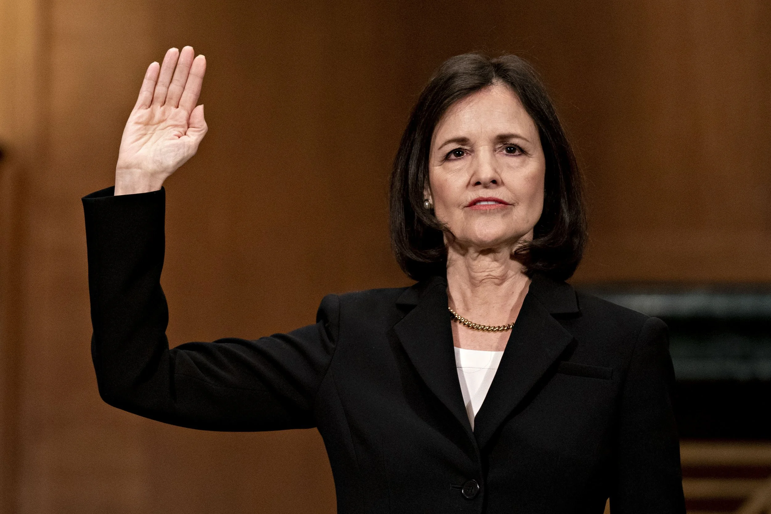 Woman in a black suit raising her right hand, possibly taking an oath, in a formal setting.