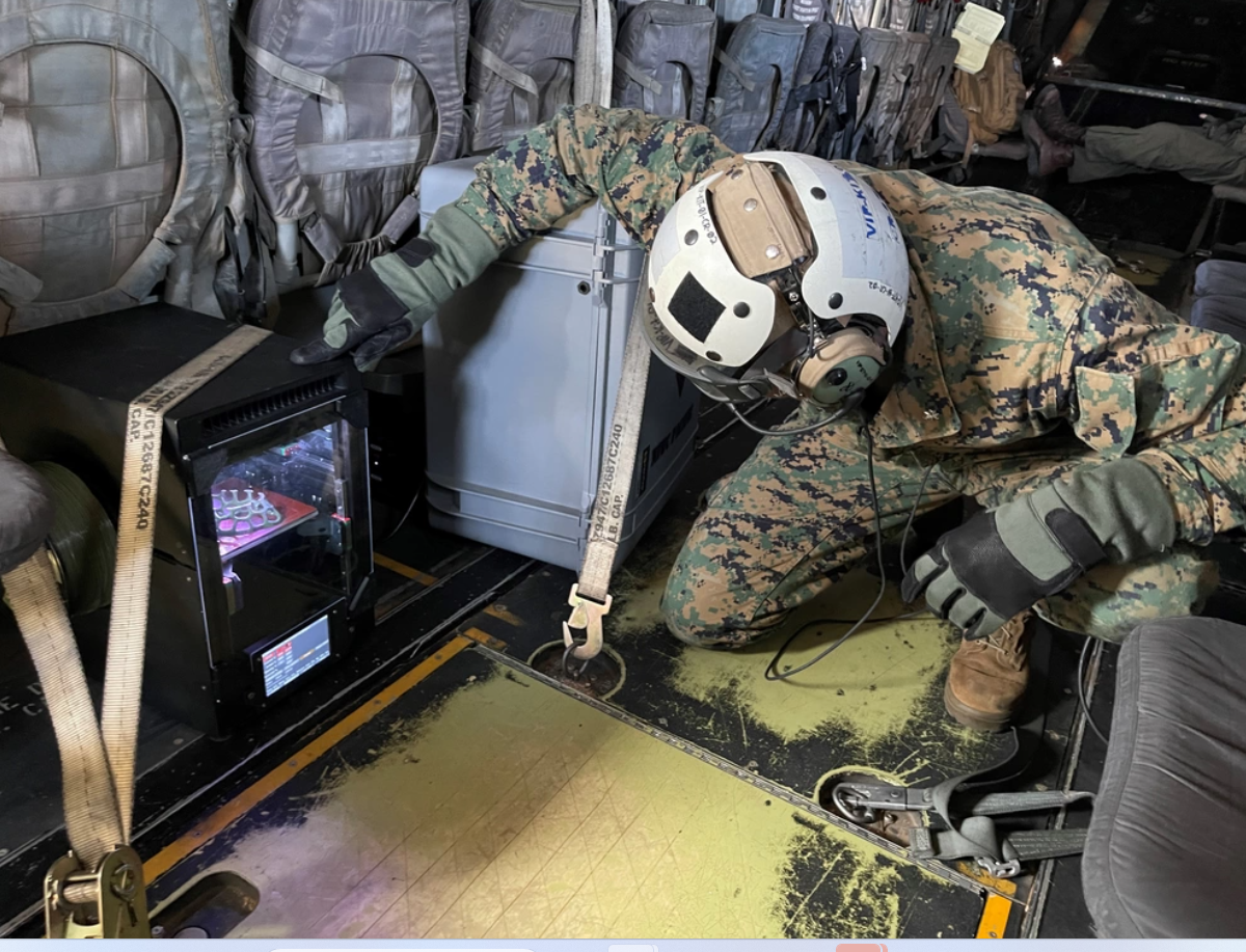 A person in military camo gear inside a helicopter, observing a 3D printer secured to the floor.