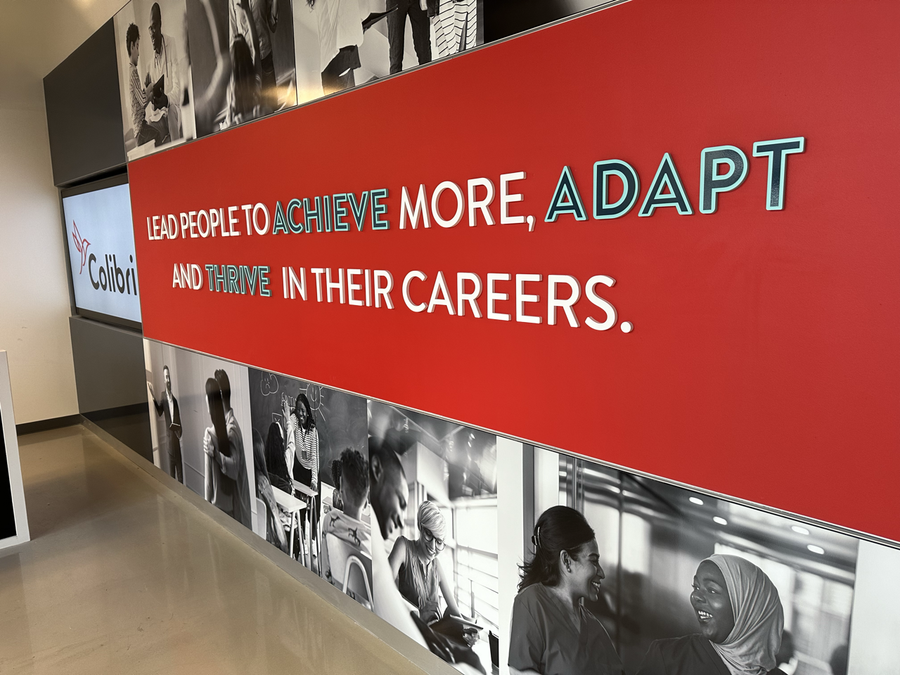 Interior wall with a red background featuring the phrase 'Lead people to achieve more, adapt and thrive in their careers' in colorful 3D lettering, with black and white photos of diverse people working and interacting
