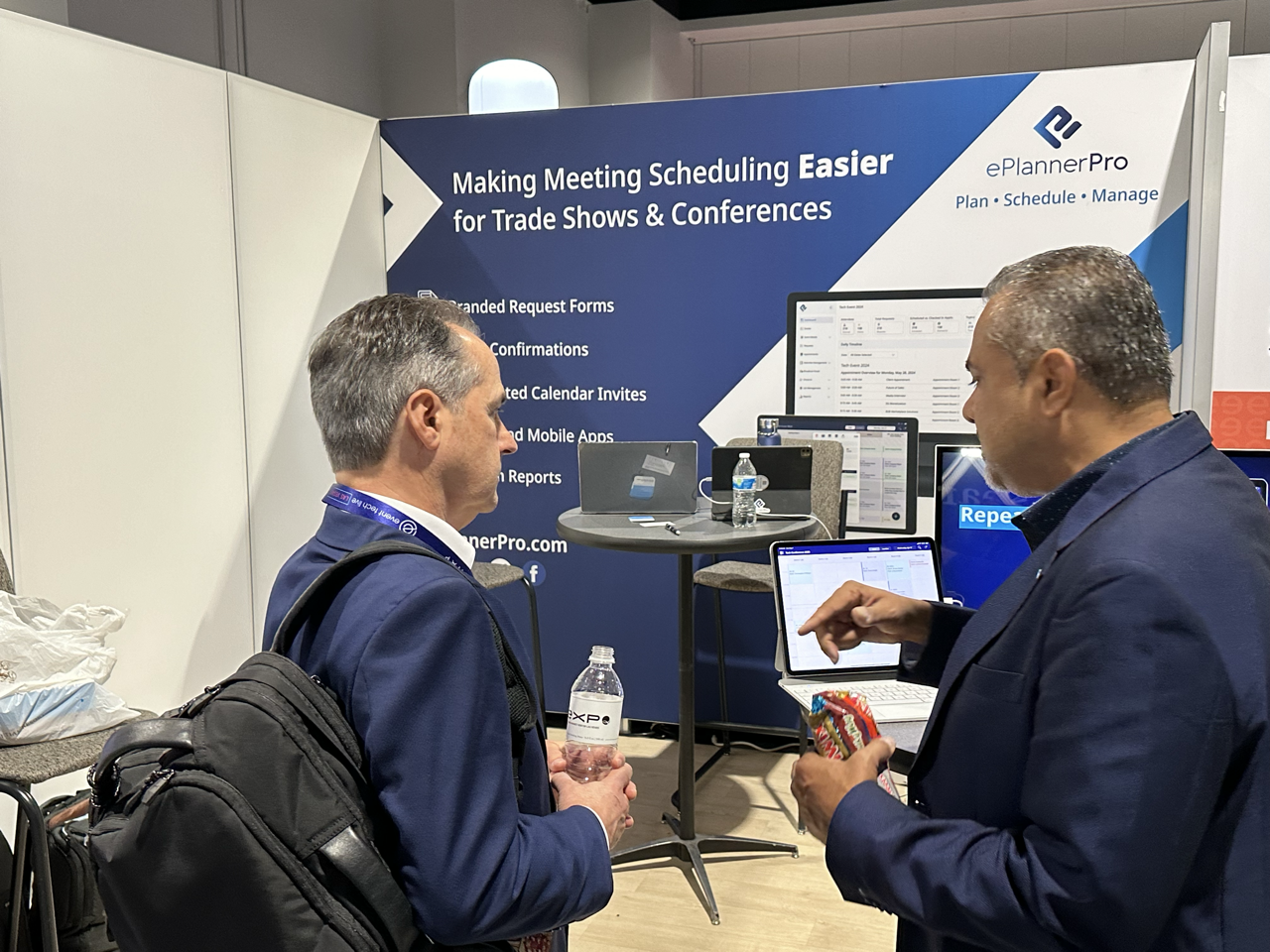 Two men in suits having a conversation in front of a booth promoting ePlannerPro, a meeting scheduling software, at a conference or trade show, with computer screens and promotional materials in the background.