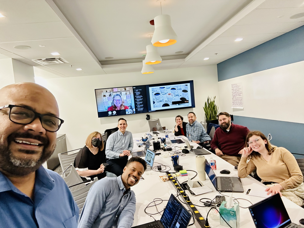 Group of eight people in a conference room, some with laptops and notebooks, smiling and posing for a photo, with a large display screen behind them showing a virtual participant.