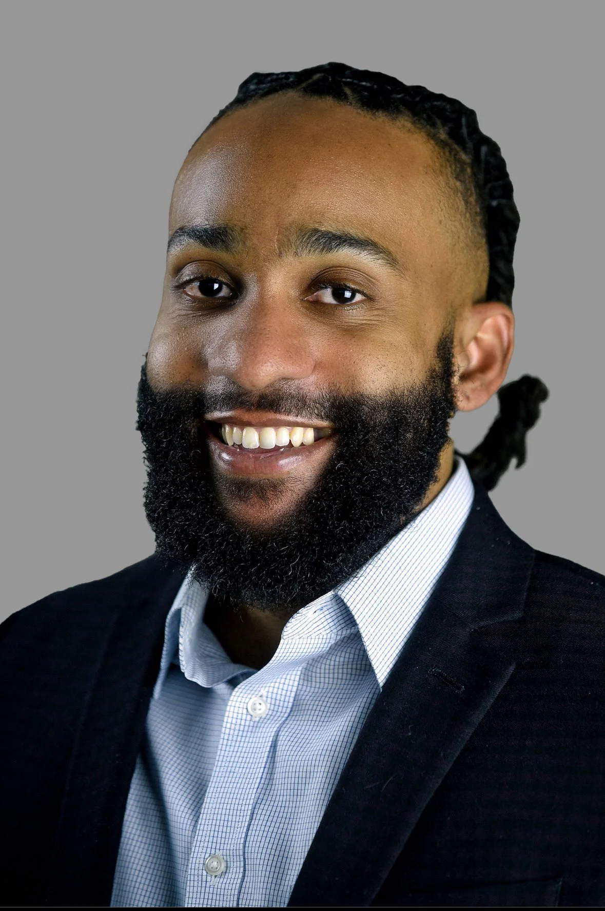 Headshot of a smiling man with a beard and dreadlocks, wearing a black blazer and a white shirt, against a gray background.