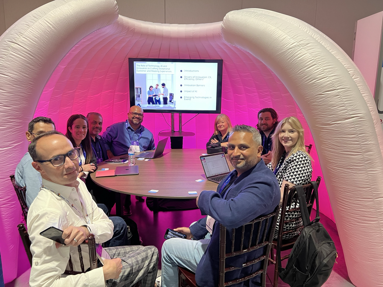 Group of nine people sitting around a round table during a conference or meeting, with a large monitor displaying a presentation behind them. The setting features a pink inflatable structure.