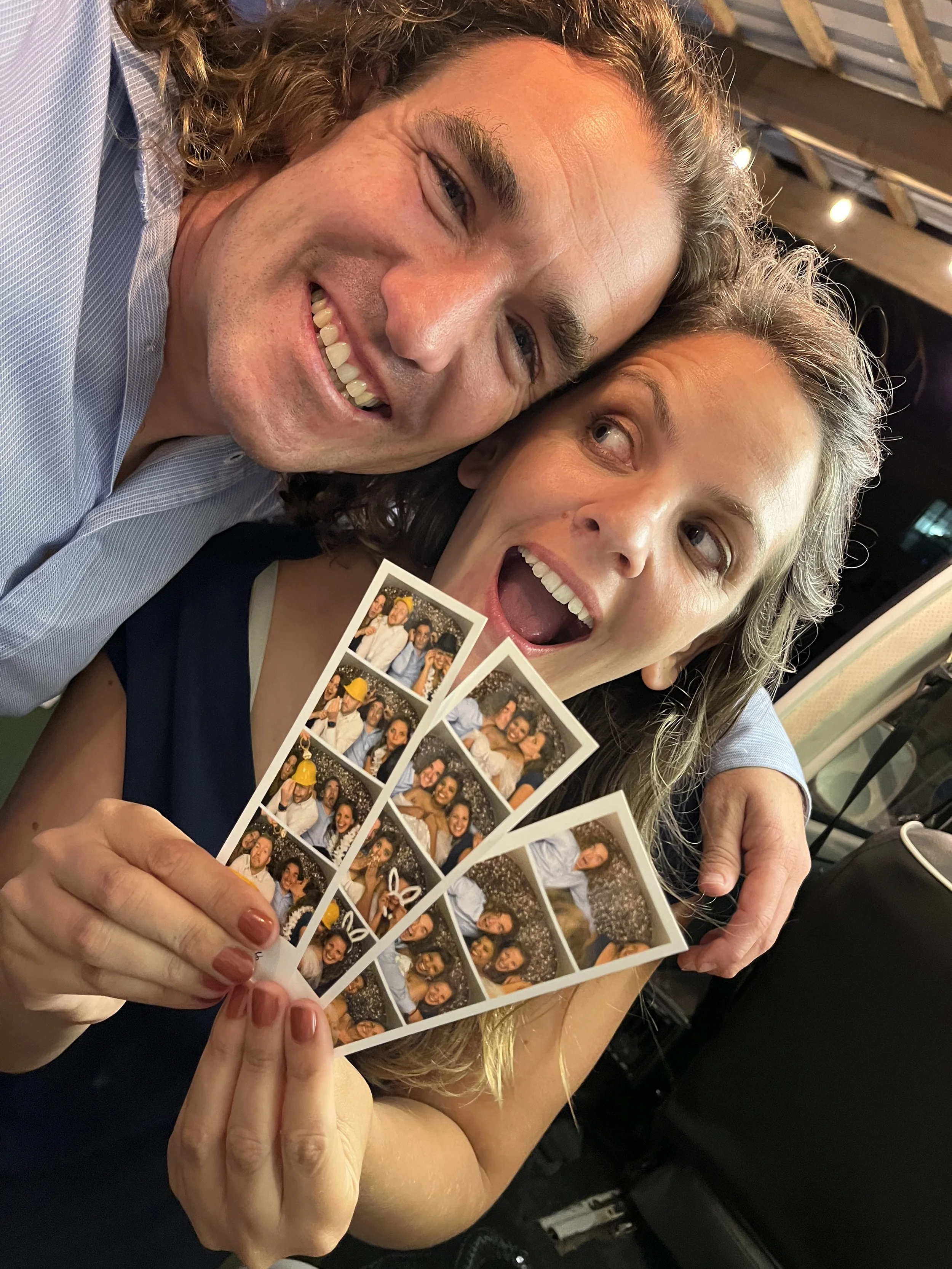 A happy man and woman celebrating together at a party, holding photo booth strips with multiple pictures, smiling, with the woman looking at the man.