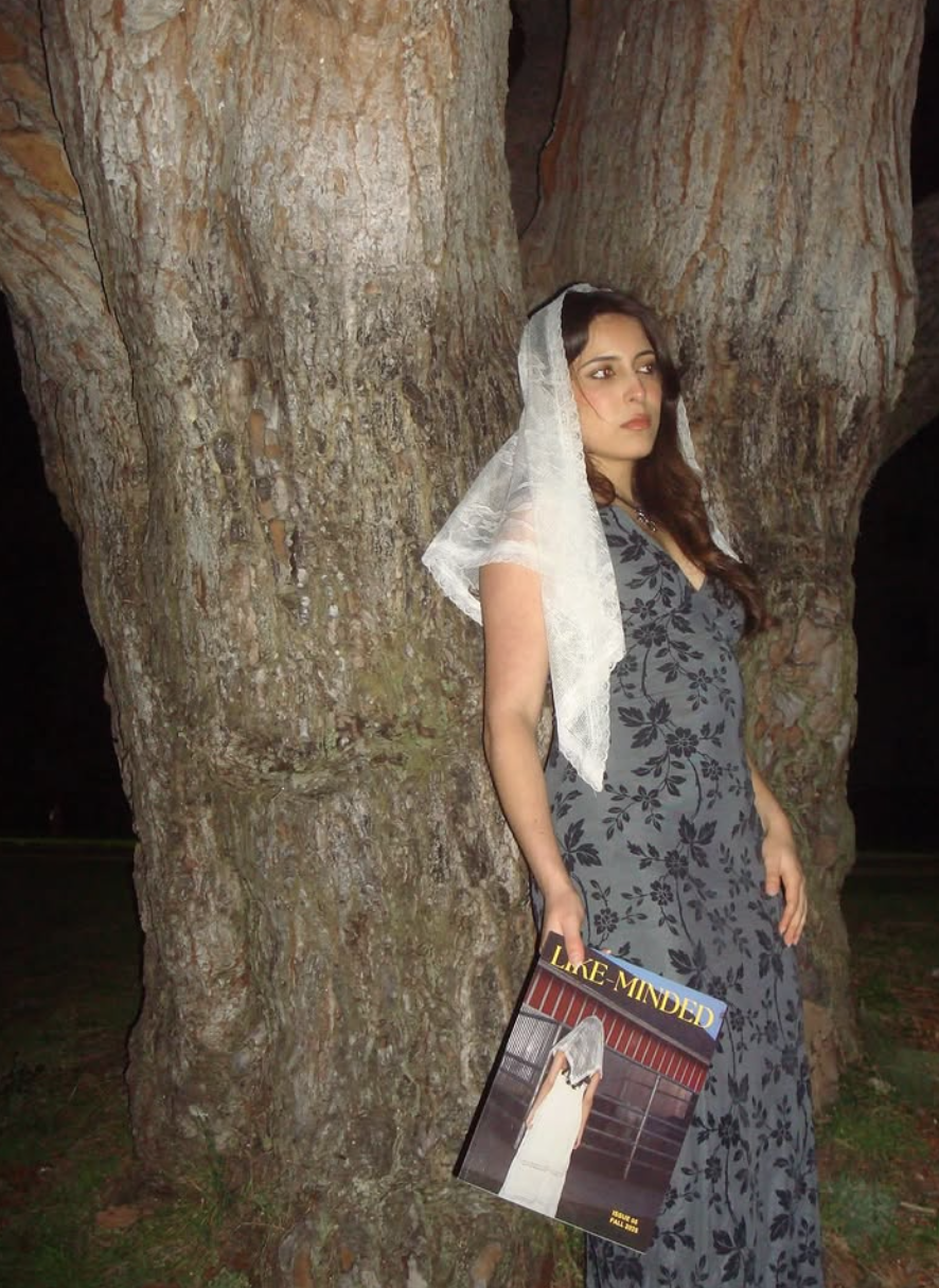 A woman in a grey dress with black floral pattern, wearing a white veil, stands against a large tree trunk at night, holding a magazine titled 'Love Mined' in her right hand.