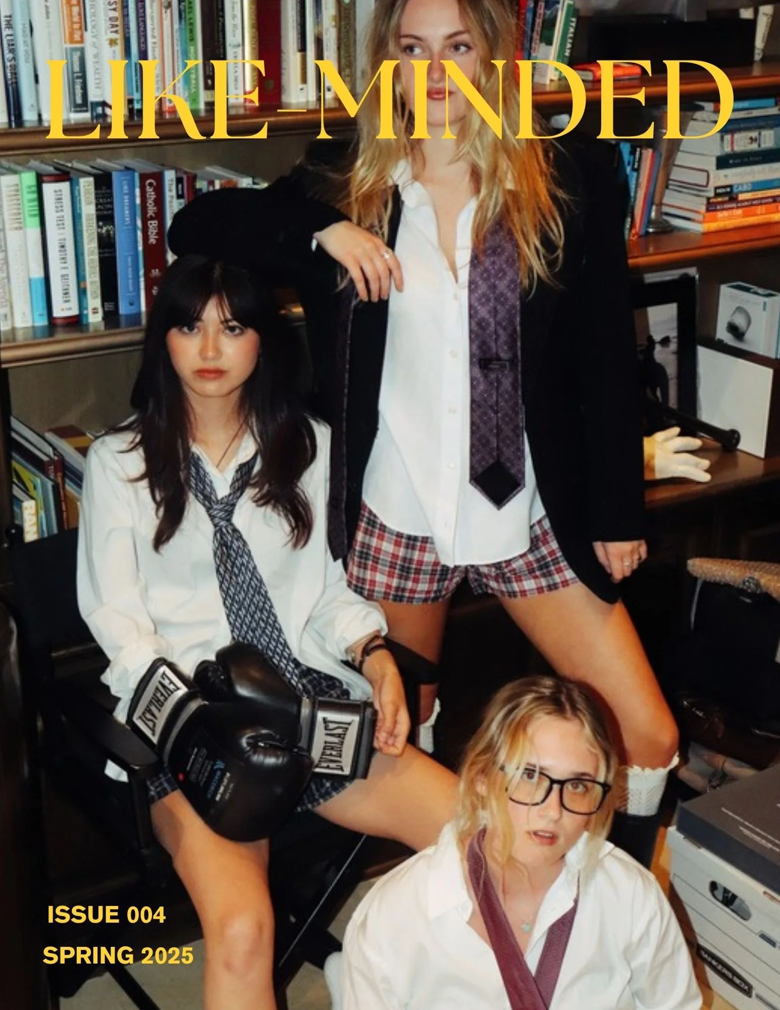 Group of young women in a library or study room, with one woman in the foreground wearing glasses and a white shirt, another woman standing with boxer gloves, and a third woman with blonde hair and glasses sitting on the floor. The background contains bookshelves filled with books and some printed materials. Overlaid on the image is the text 'LIKE - MINDED' in large yellow font, along with additional text 'ISSUE 004 SPRING 2025' in the bottom left corner.