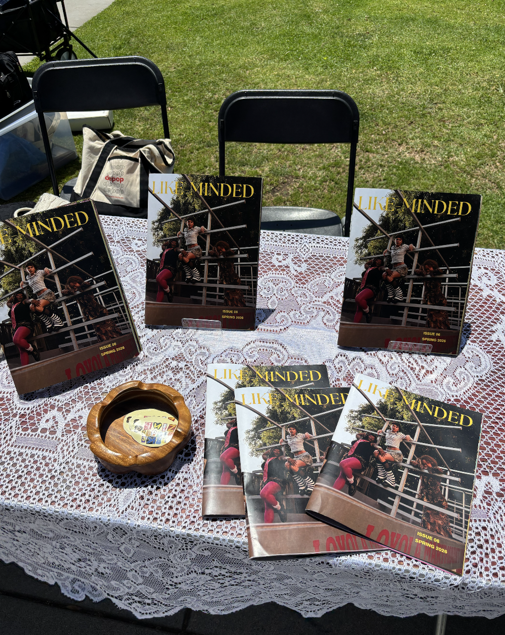 Table covered with a white lace tablecloth displaying copies of a magazine titled 'LIKE MINDED,' featuring a photo of three people near a playground structure. There is a wooden ashtray with small items inside, and behind the table, two black chairs are placed on grass.
