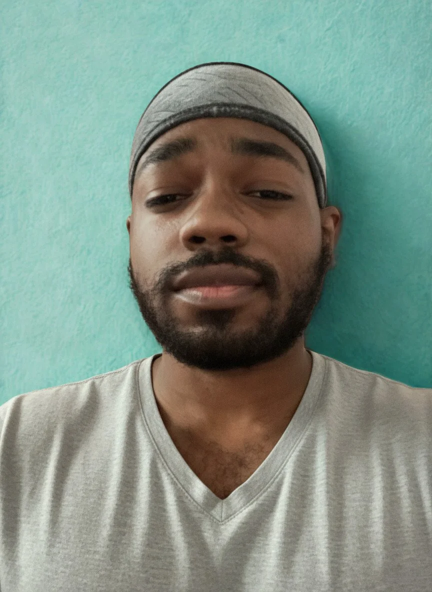A young man with a beard wearing a gray V-neck T-shirt and a gray headband, standing in front of a light teal wall.