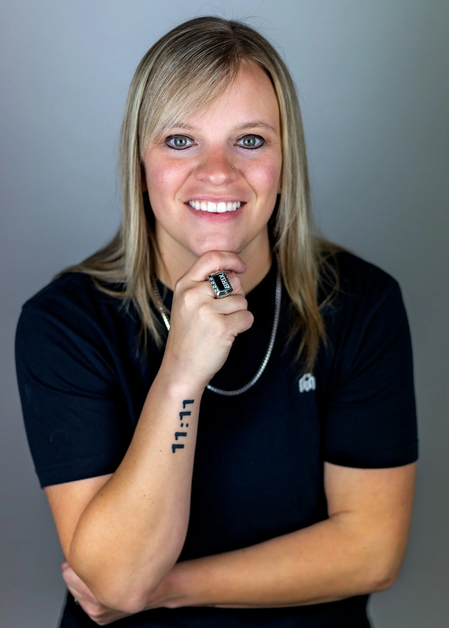 A smiling woman with blond hair and blue eyes, wearing a black t-shirt and jewelry, resting her chin on her hand with a tattoo that reads '11:11' on her forearm, against a neutral background.