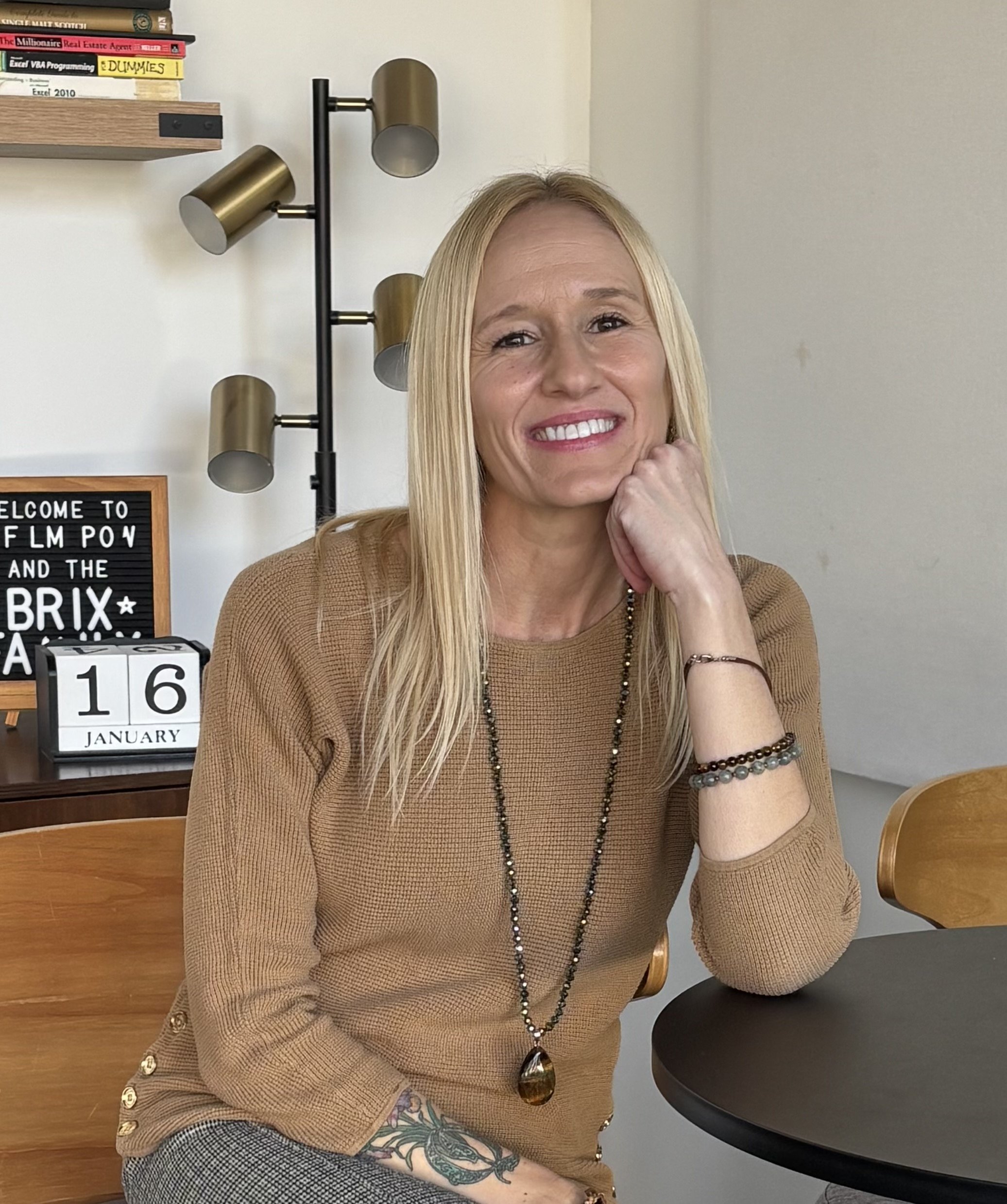 A woman with long blonde hair wearing a brown sweater and jewelry, sitting at a black round table, smiling with her head resting on her hand, in a room with a wooden chair, a bookshelf, a black letter board, a desk calendar showing January 16, and a modern floor lamp with gold and black finishes.