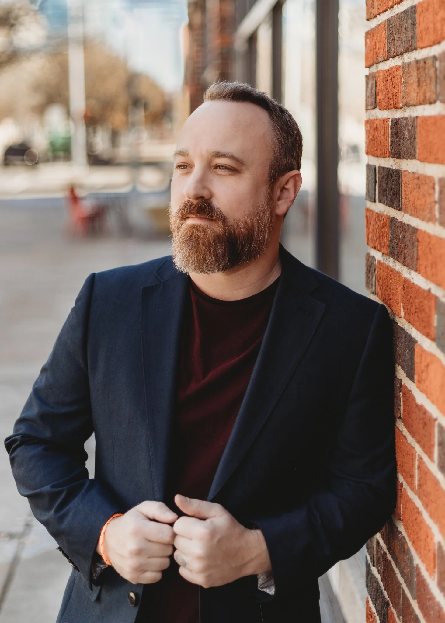 A man with a beard and short hair leaning against a brick wall, wearing a dark blazer and a dark shirt, outdoors during daytime.