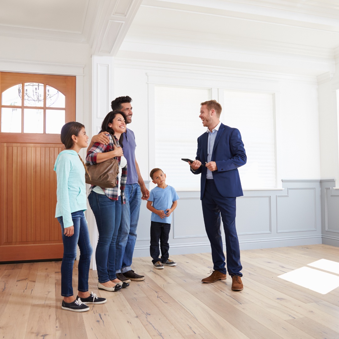A family of four, including a woman, man, girl, and boy, are smiling and talking to a real estate agent in a bright, empty room with white walls and wooden floors, likely during a house tour or purchase.