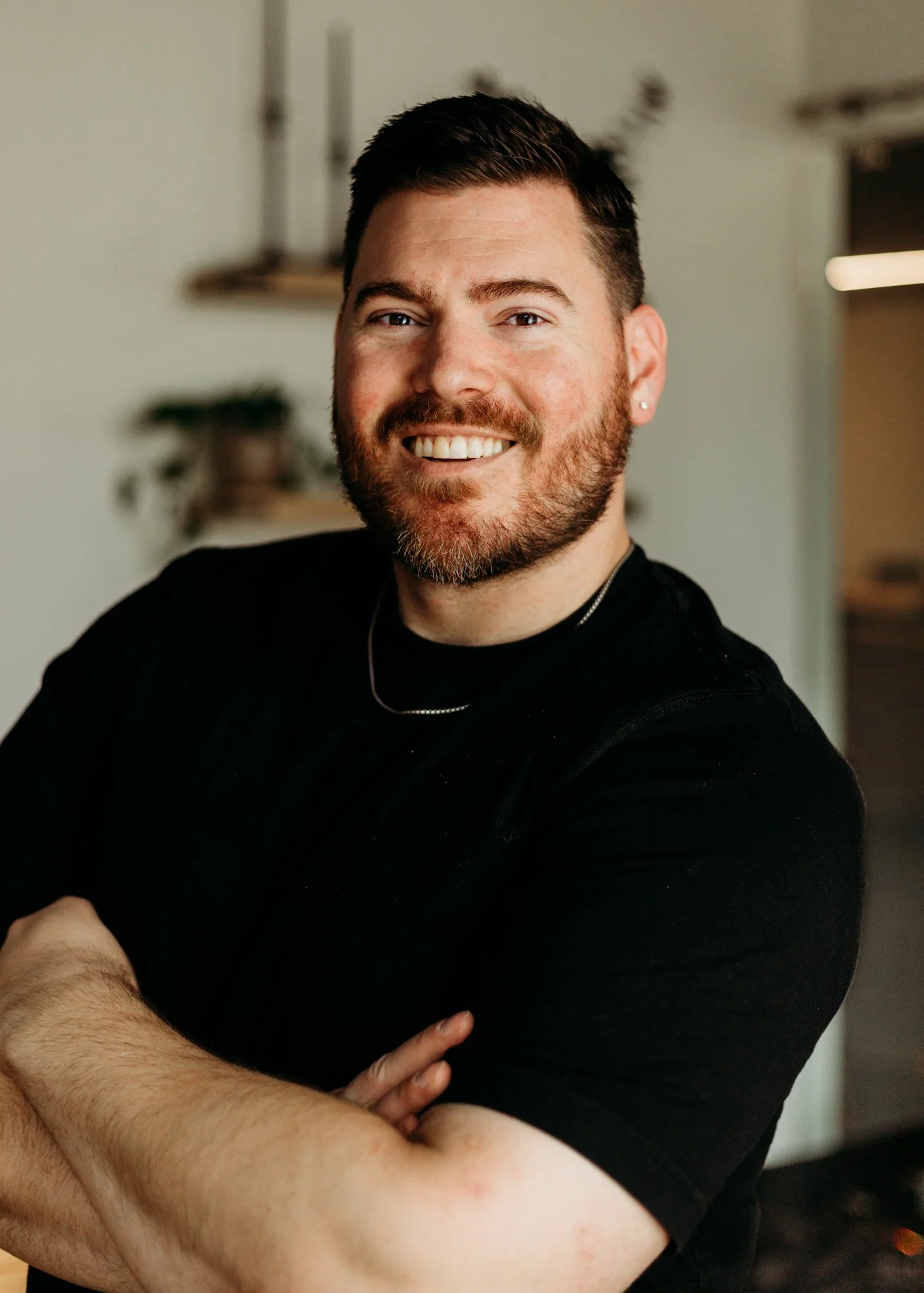 A smiling man with a beard, wearing a black shirt, standing indoors with his arms crossed.