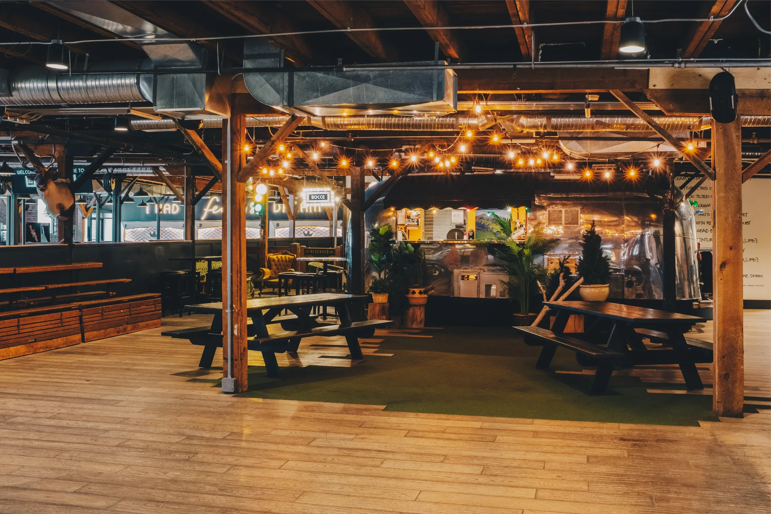 Interior of a rustic restaurant with wooden beams, string lights, wooden picnic tables, potted plants, and a metallic food truck at the back.