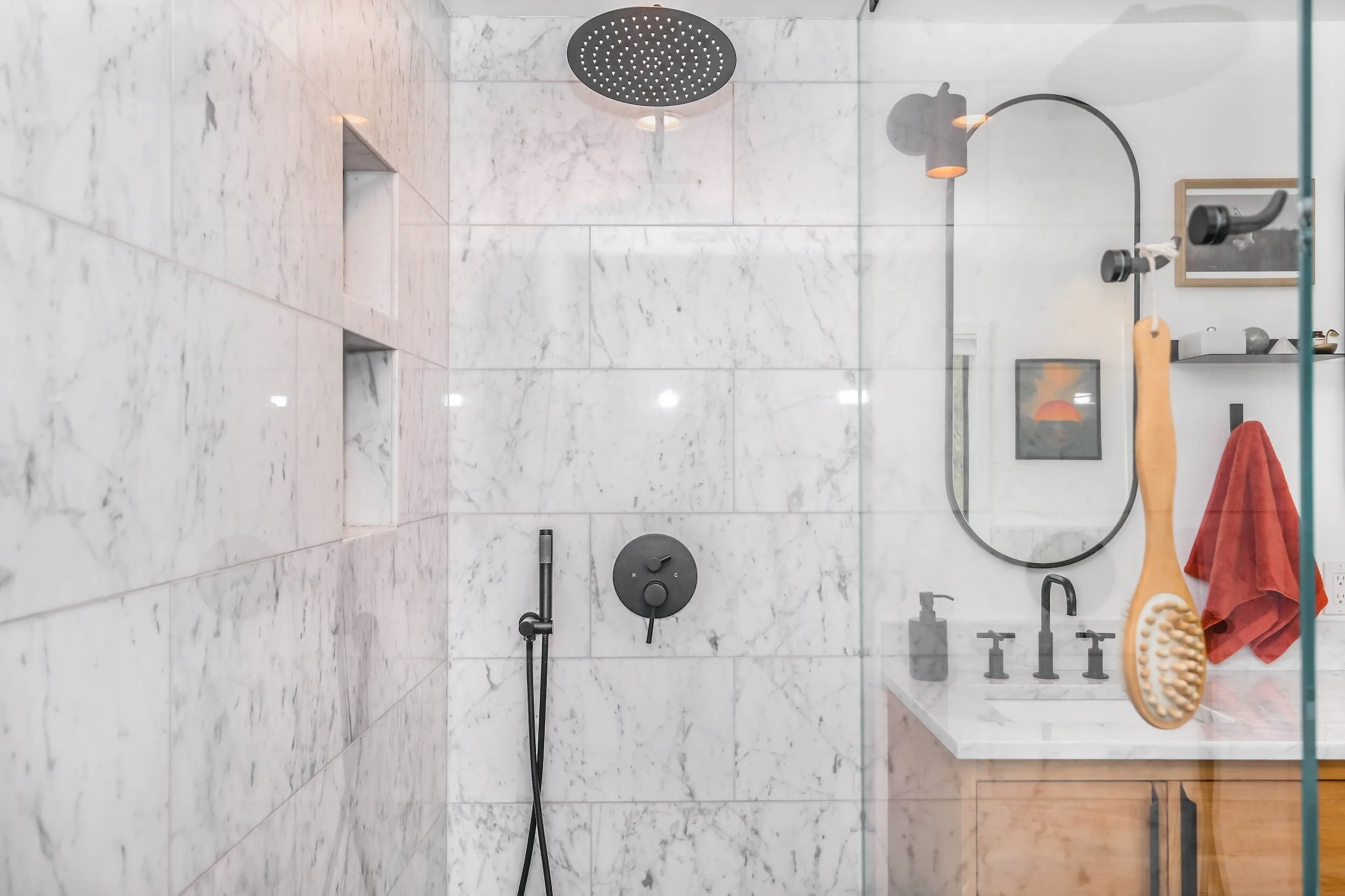 Modern bathroom featuring a marble-tiled shower with a rain shower head, black fixtures, oval mirror, wooden vanity, and a red towel.