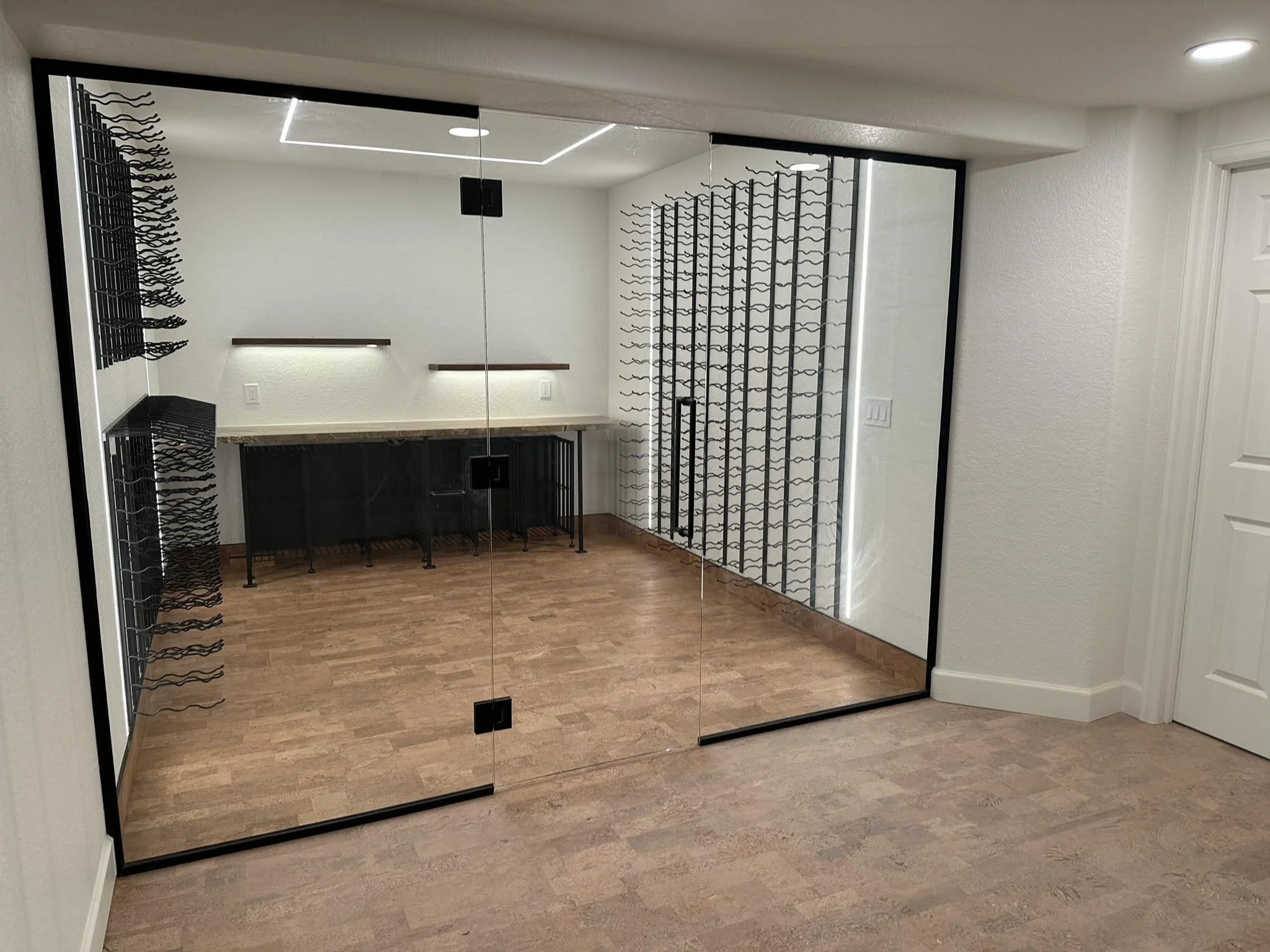 Modern wine room with glass doors, empty black metal wine racks on the walls, light-colored wooden floor, and minimalistic lighting.