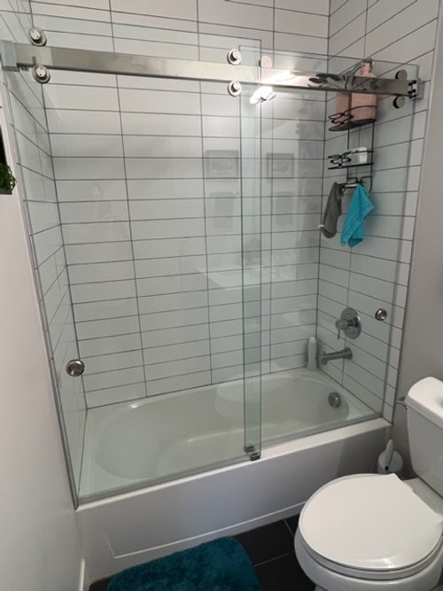 Bathroom with white tiled shower, glass sliding door, bathtub, towel rack, toilet, and blue mat on the floor.