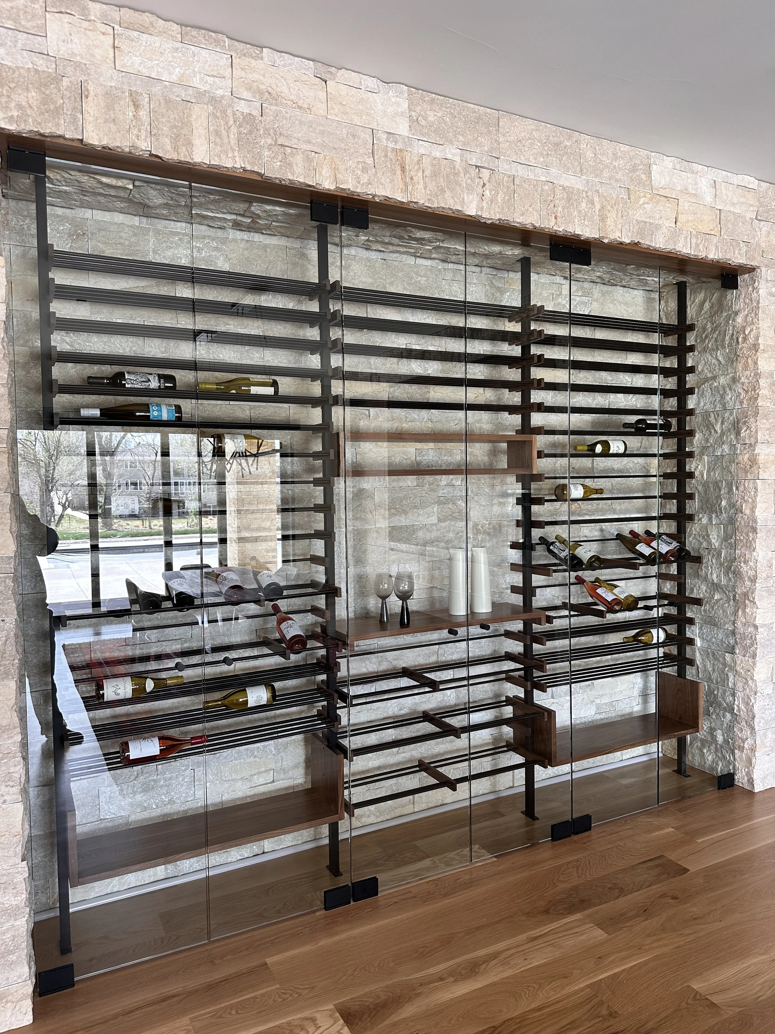 Glass-enclosed wine rack with wooden shelves inside a stone wall, holding various wine bottles and wine glasses on display.