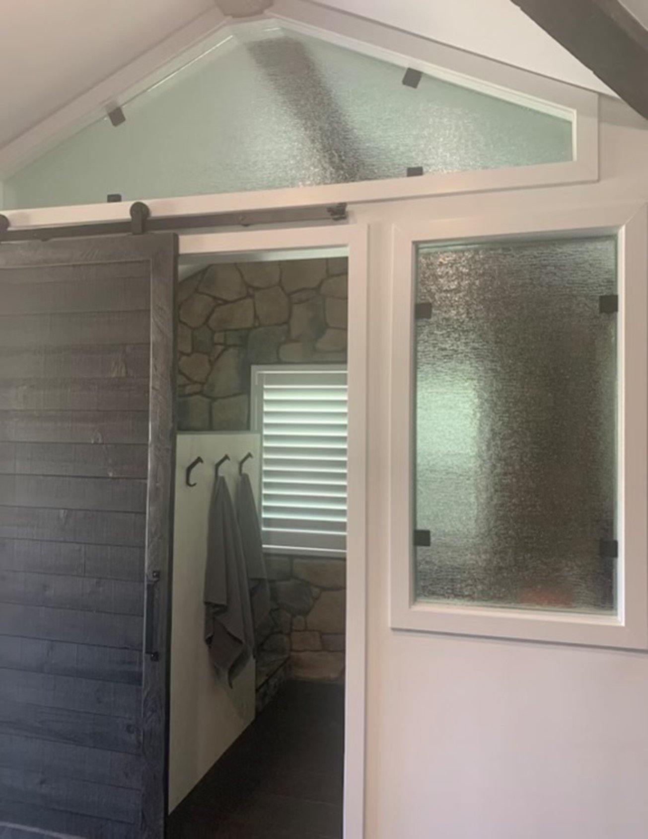 Interior view of a rustic-style bathroom with a sliding wooden barn door and stone wall. The bathroom features frosted glass windows, hooks on the wall with gray towels, and a window with closed blinds.
