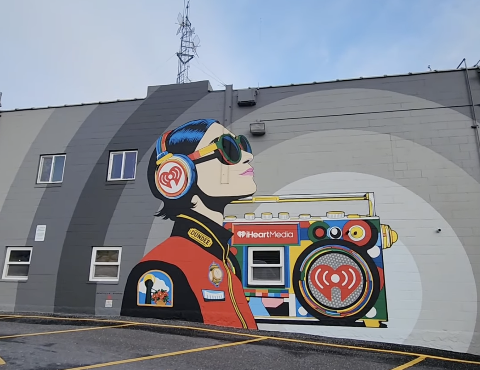 Colorful mural of a woman with dark hair and sunglasses, holding a boombox with the iHeartMedia logo, on the side of a building with small windows and a gray background.