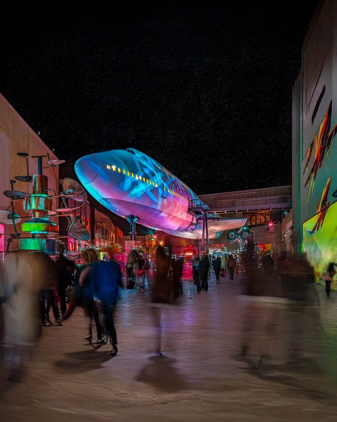 Night view of a space-themed exhibit with a large spaceship model on display, colorful lighting, and visitors walking around in the foreground.