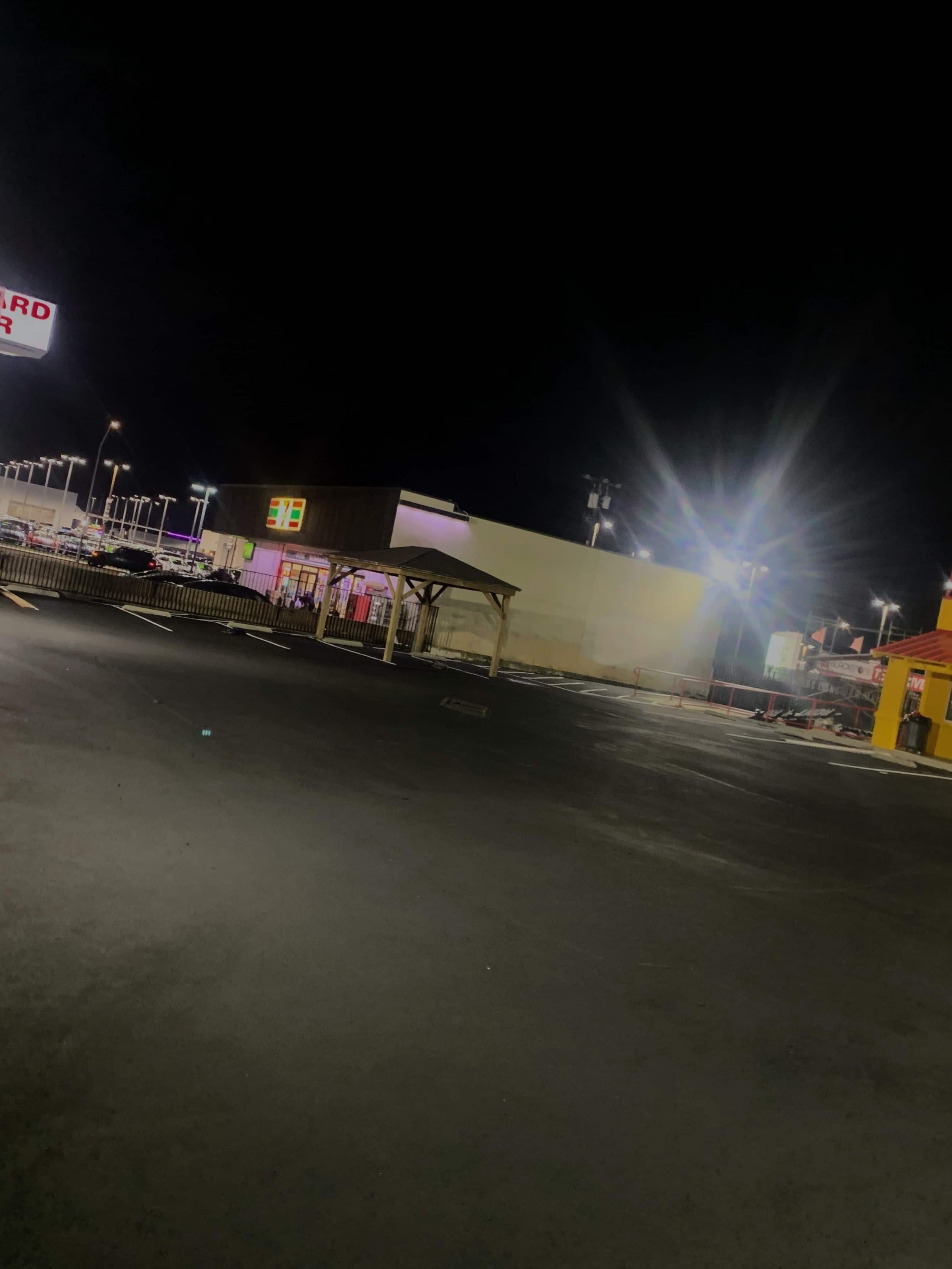 An empty parking lot with fresh pavement sealing at night, a brightly lit 7-Eleven store shining under the dark sky.