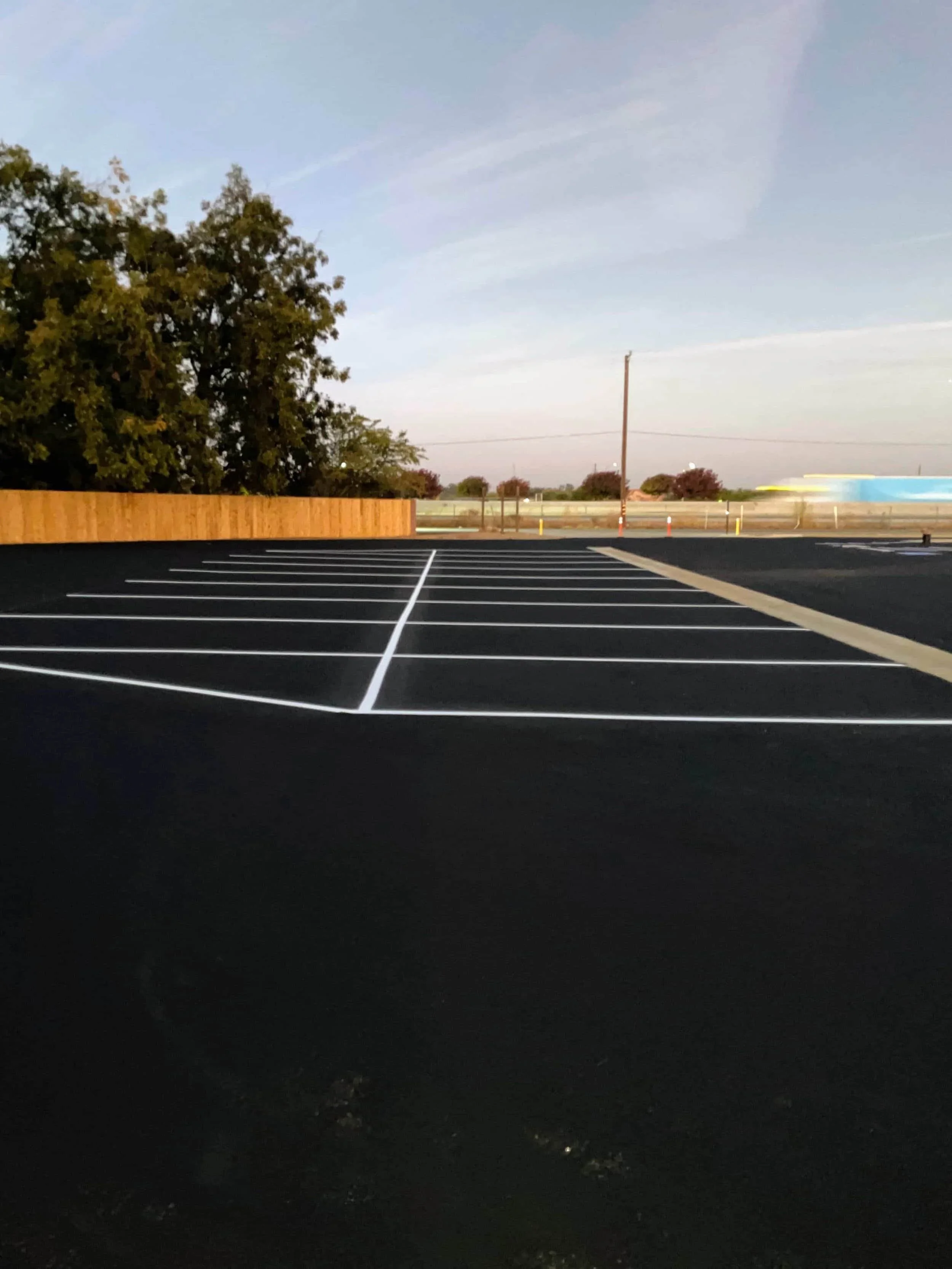 Empty parking lot with freshly painted white lines after sealcoating Fresno, a wooden fence, and trees in the background at dusk.