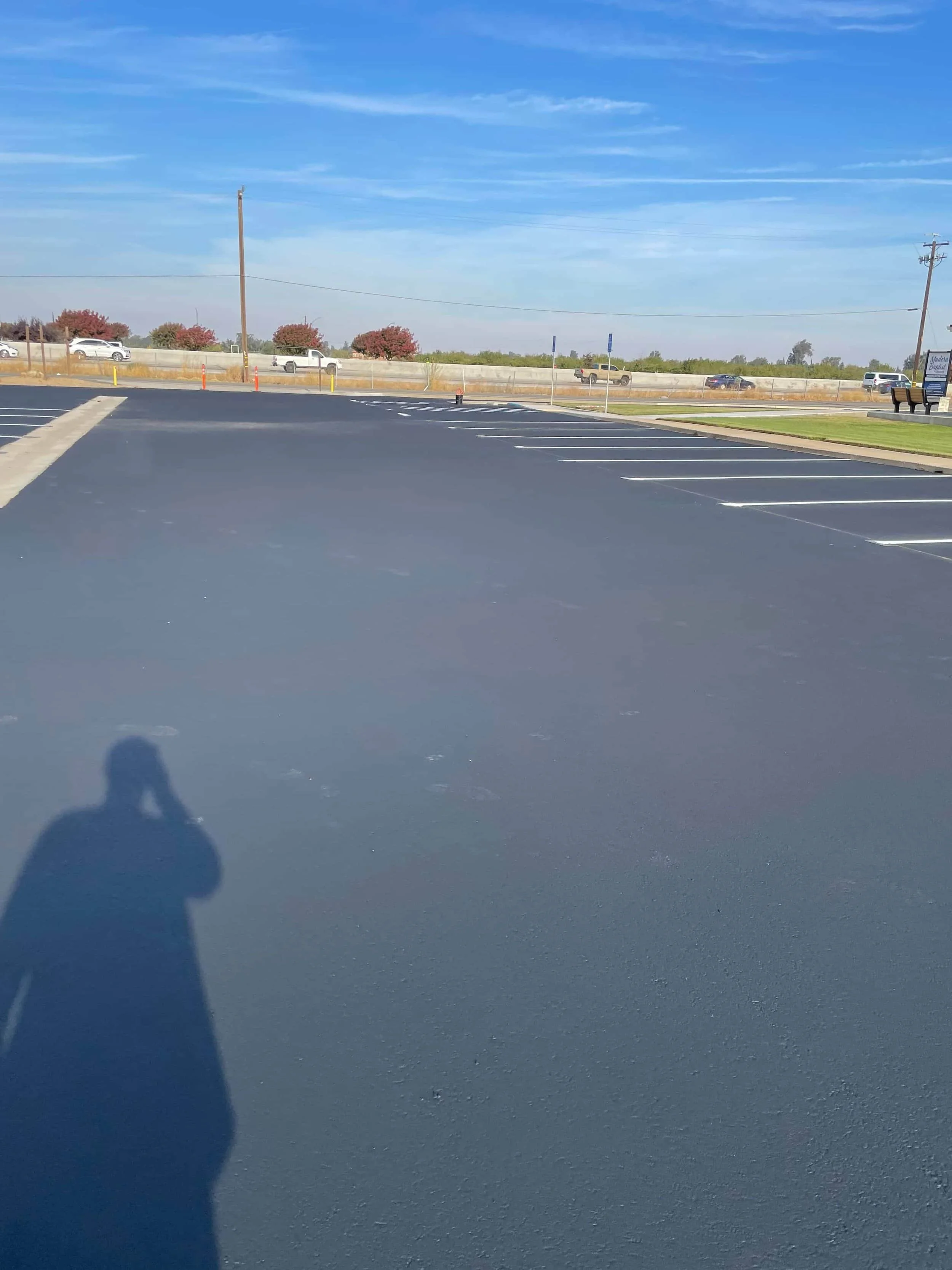 A parking lot with freshly paved black asphalt, a small landscaped island with a tree and bushes, and a building in the background under a clear blue sky - asphalt paving fresno.