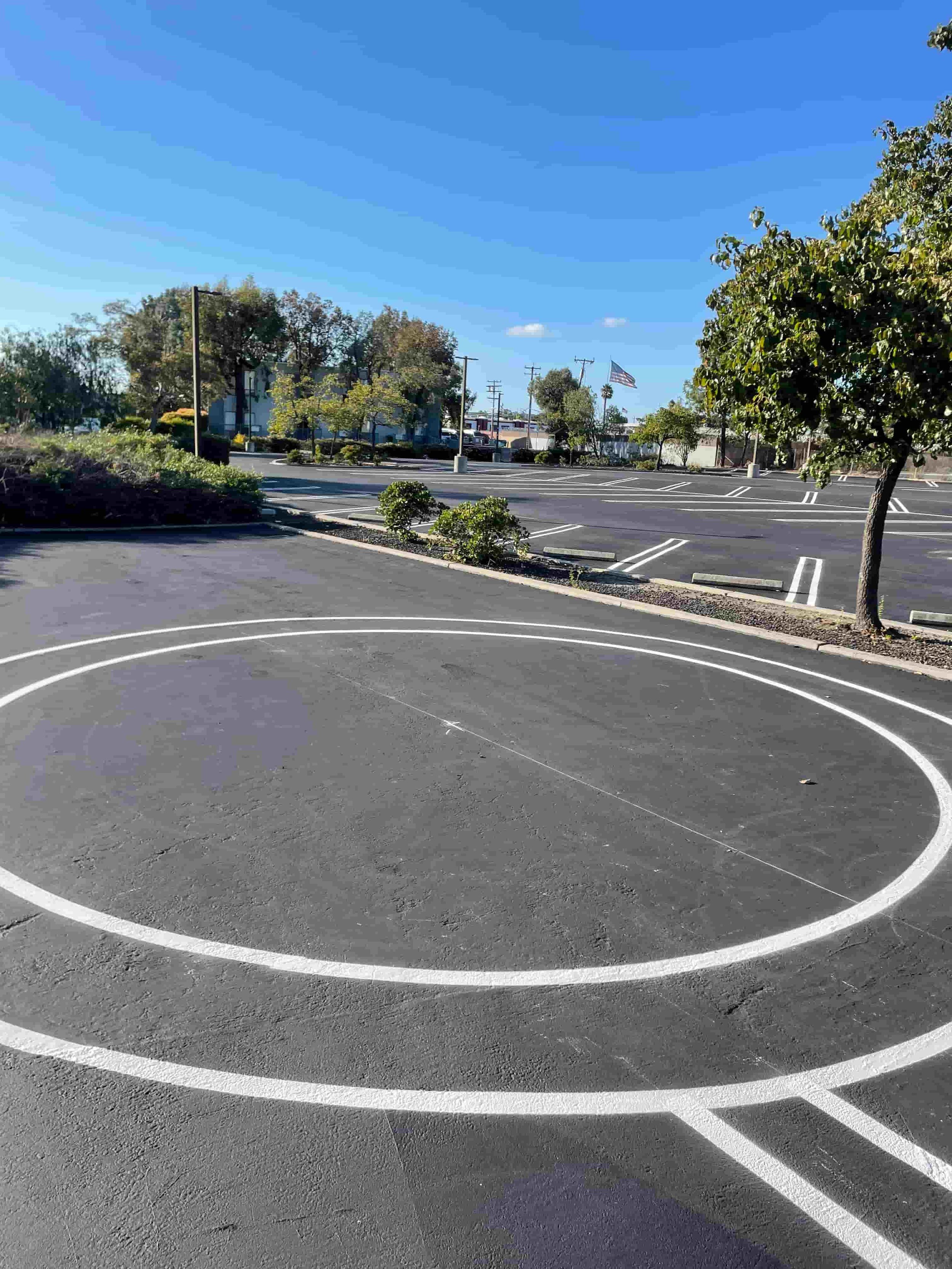 Freshly painted white lane lines curve on a paved, empty parking lot under a clear blue sky, with trees and parked cars in the distance.