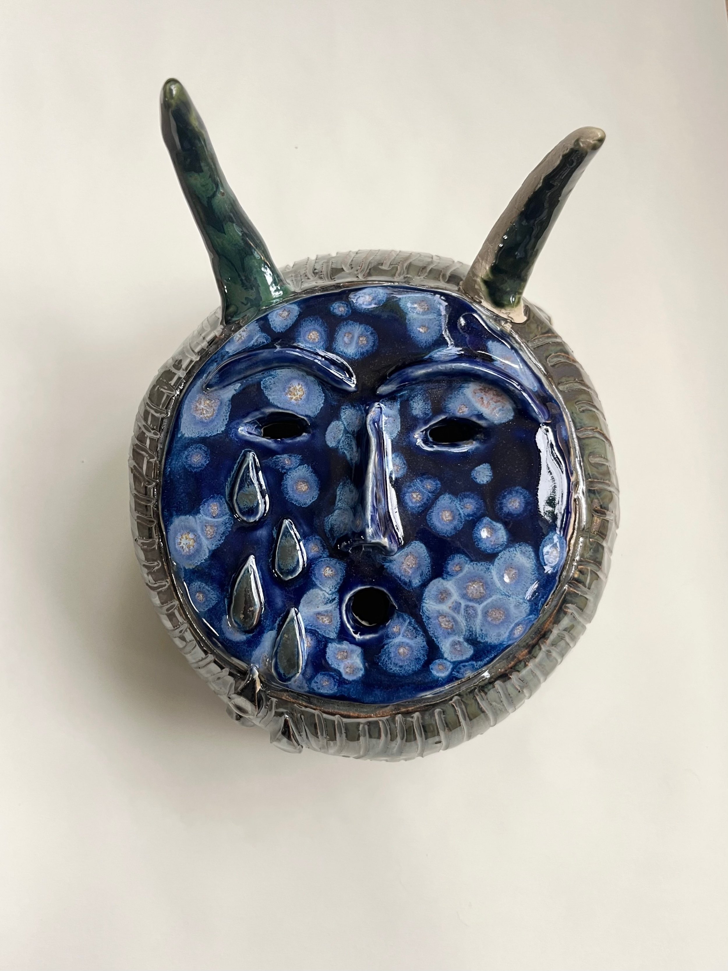 Glass art piece depicting a blue face with closed eyes, teardrops, and floral patterns, with two green and gray antennae-like extensions.