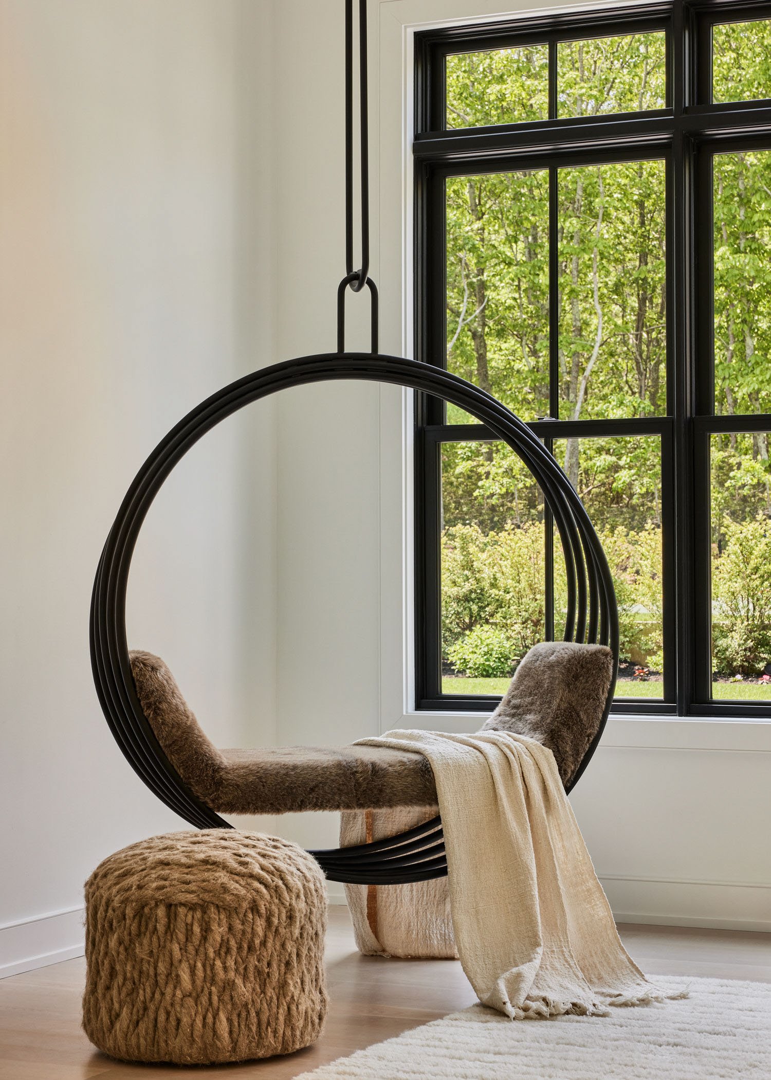 Modern interior with a black circular hanging chair, beige blanket, and brown pouf beside large window with a view of greenery.