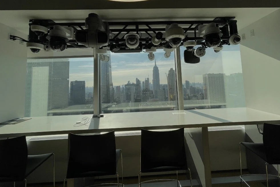 View of a city skyline through large windows in a room with white walls, a long white table, and black chairs, with a ceiling-mounted array of security cameras and equipment.