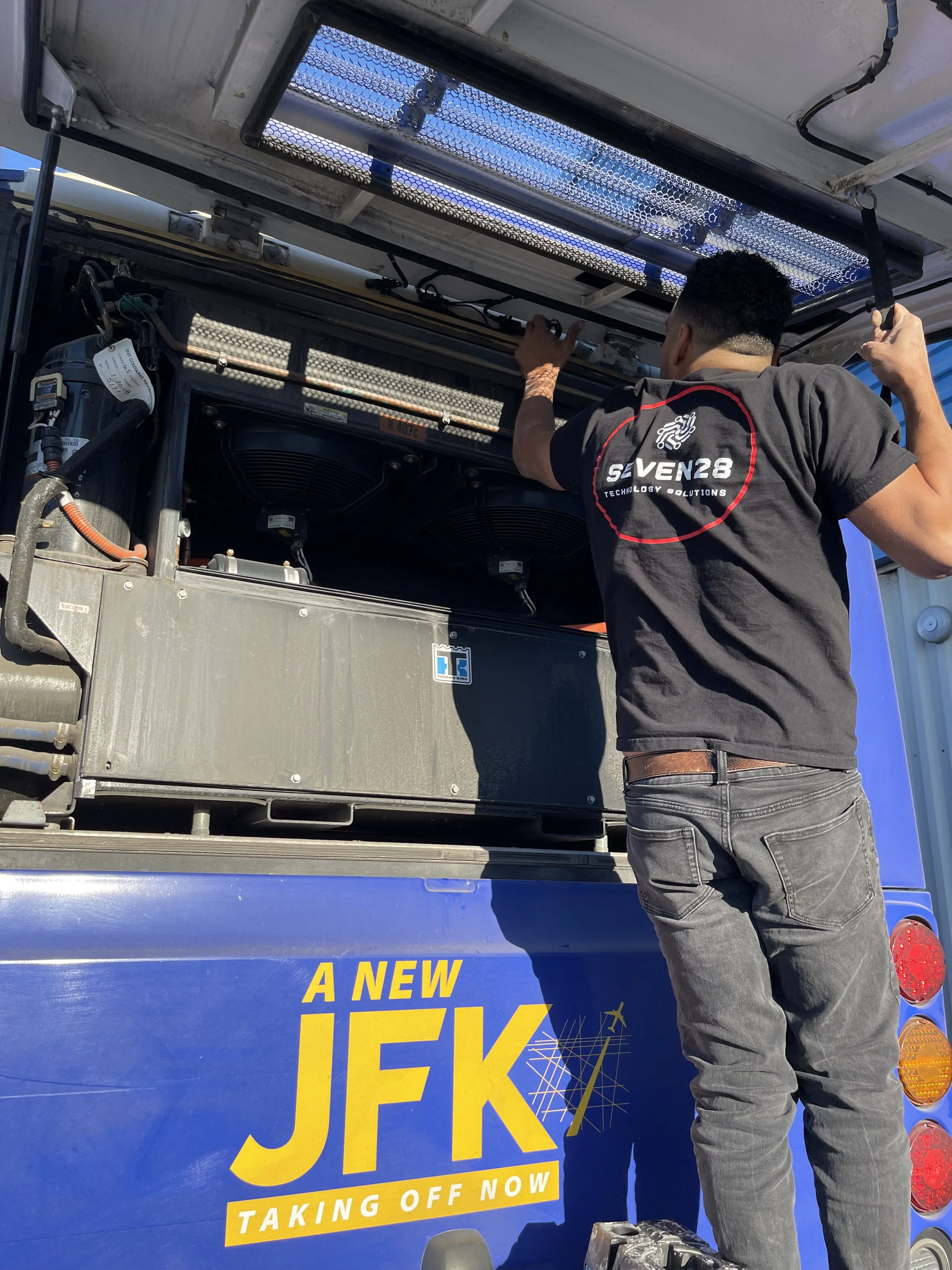 A person is working on the engine of a vehicle. The person is standing with their back to the camera and is wearing a gray t-shirt with a logo that reads 'GIVEN28 TECHNOLOGY SOLUTIONS'. The vehicle has a blue exterior with yellow text and is advertis