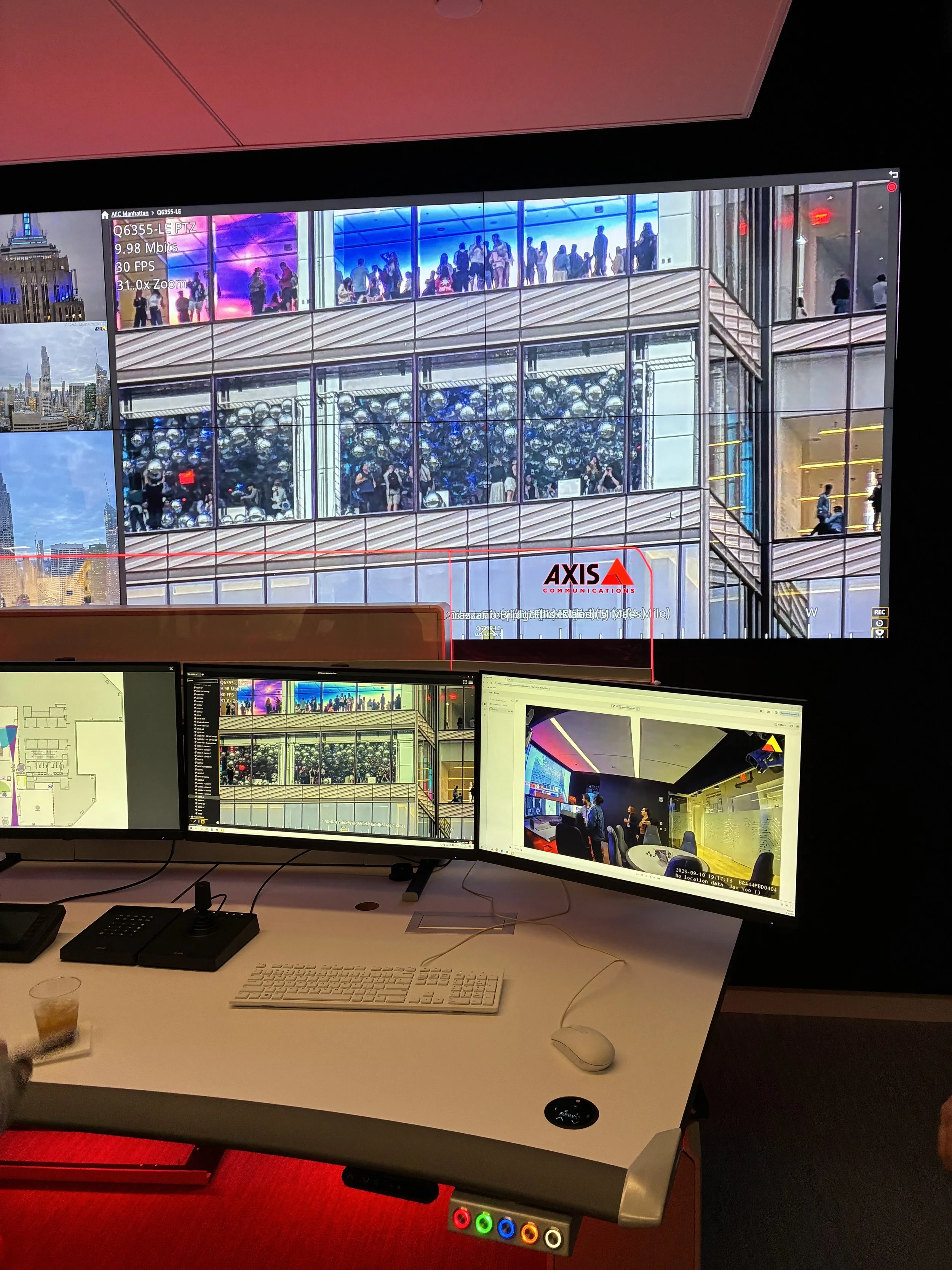 Office desk with three monitors displaying building interiors and urban landscapes, keyboard, mouse, and a glass of iced tea on a white desk, with a red light-emitting device underneath.