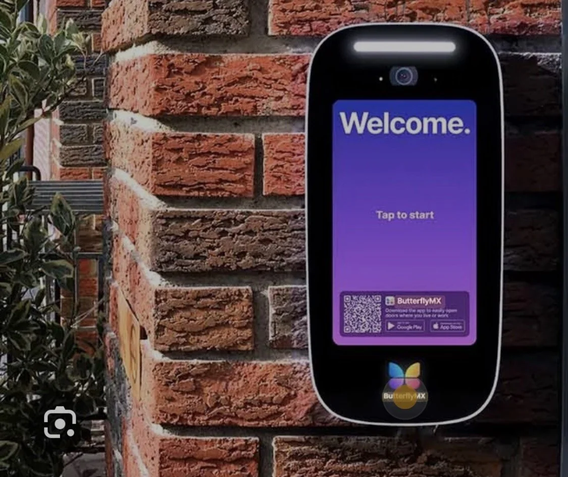 A digital sign with a purple screen displaying 'Welcome' and 'Tap to start,' mounted on a brick wall outside. The sign promotes the ButterflyMX app, with a QR code and download links for Google Play and the App Store.