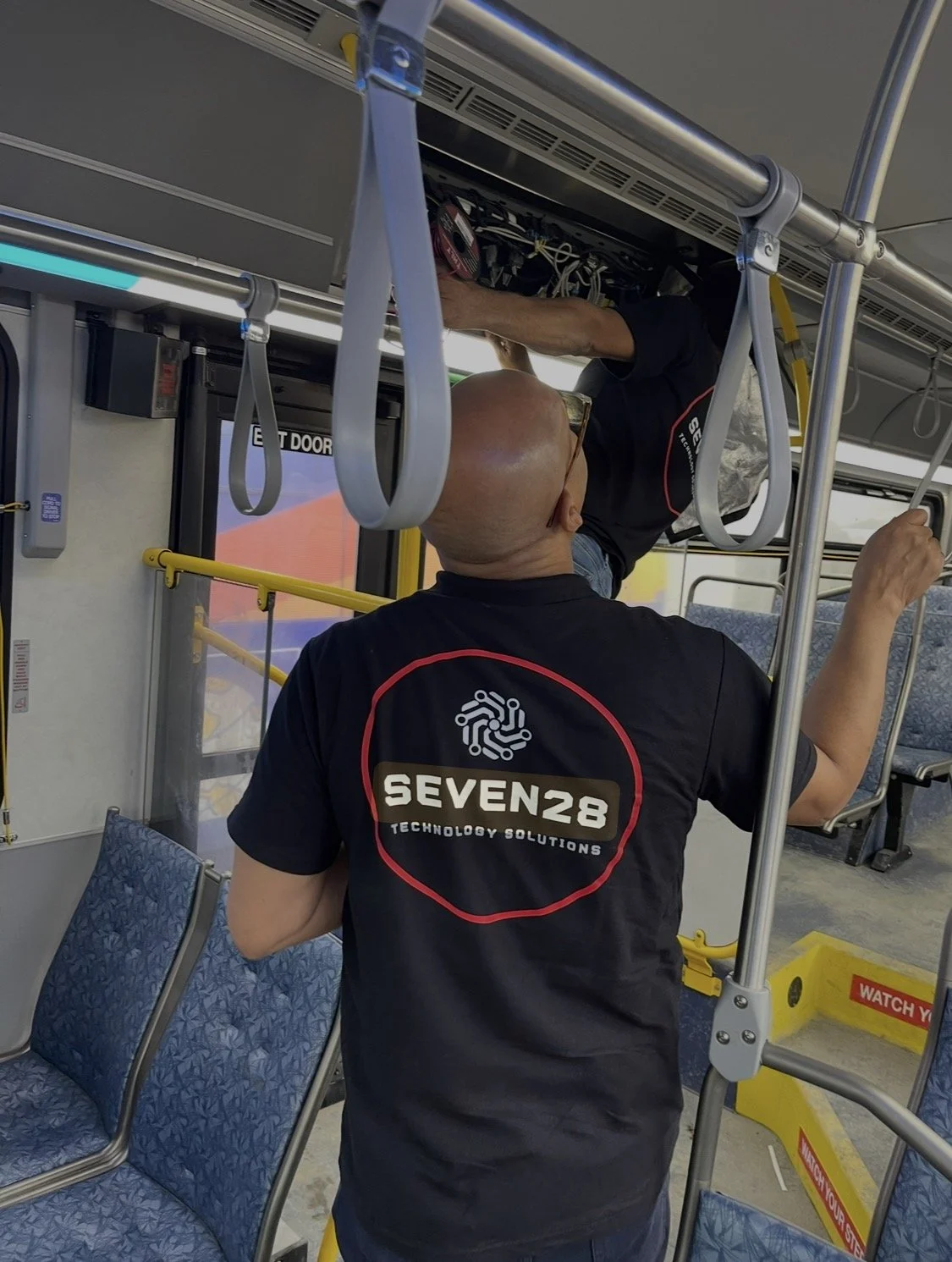 Two men wearing black shirts with 'SEVEN28 TECHNOLOGY SOLUTIONS' logo, one with a shaved head, are working on electronic equipment above their heads on a bus, with seats and yellow safety signs visible.
