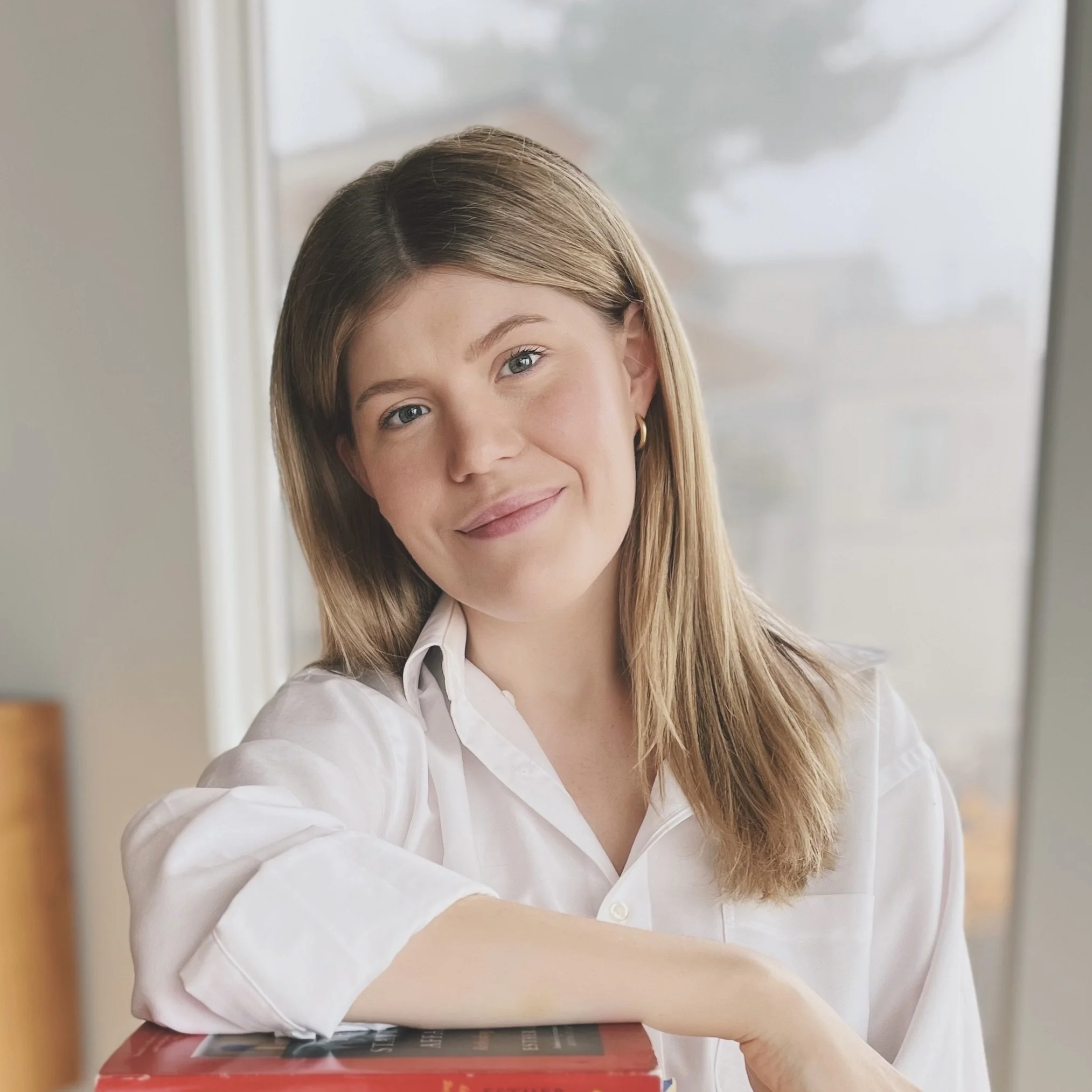 Close-up of a woman therapist with light brown hair, blue eyes, and fair skin with a warm expression, wearing a white collared shirt, sitting at a table with a book or magazine in front of her, in a bright room with large windows.