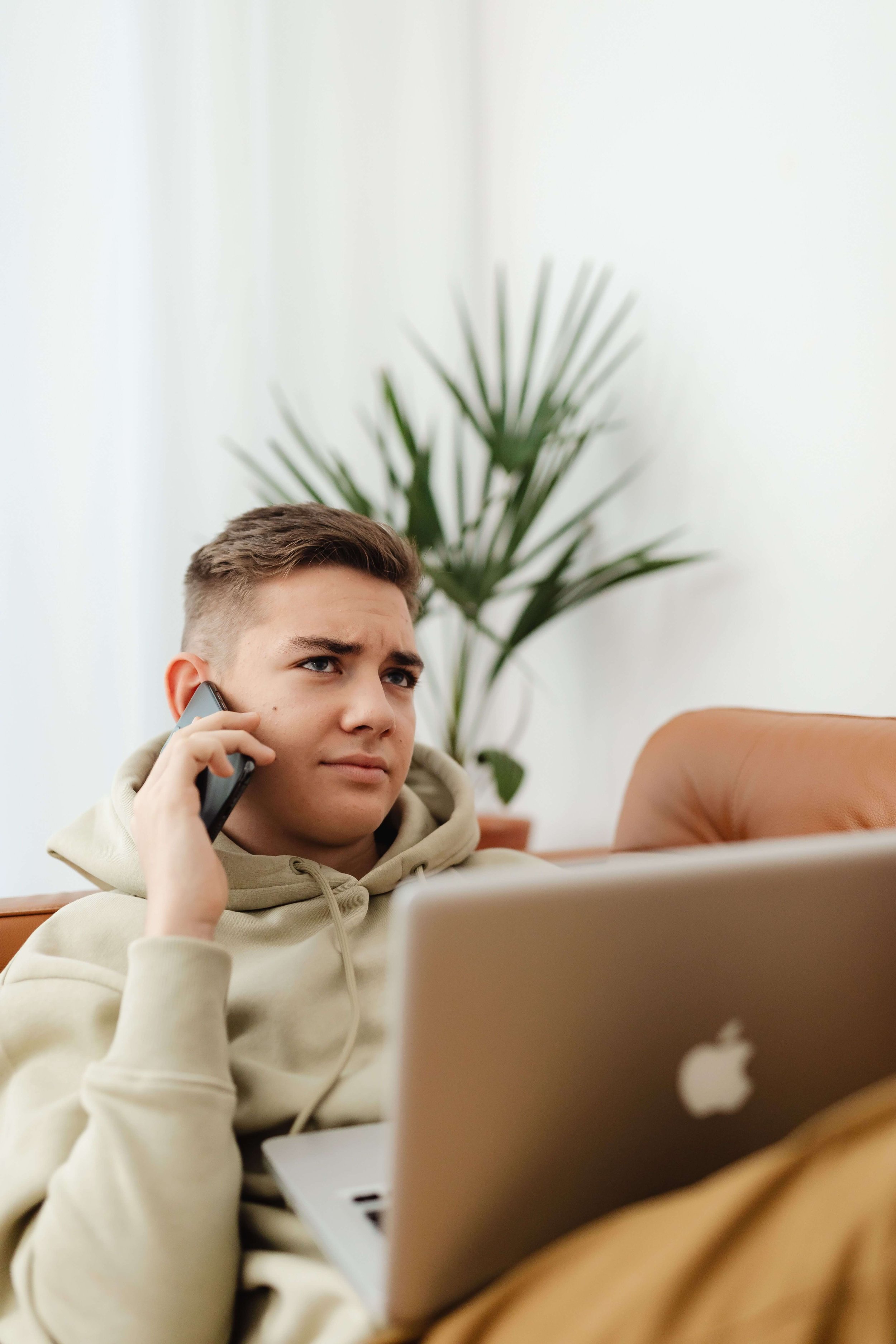 A teen boy scrunching his eyebrows while holding a phone to his ear & looking at laptop. Teen counseling in Miami, FL can help your teen manage their emotions better. Learn more here.