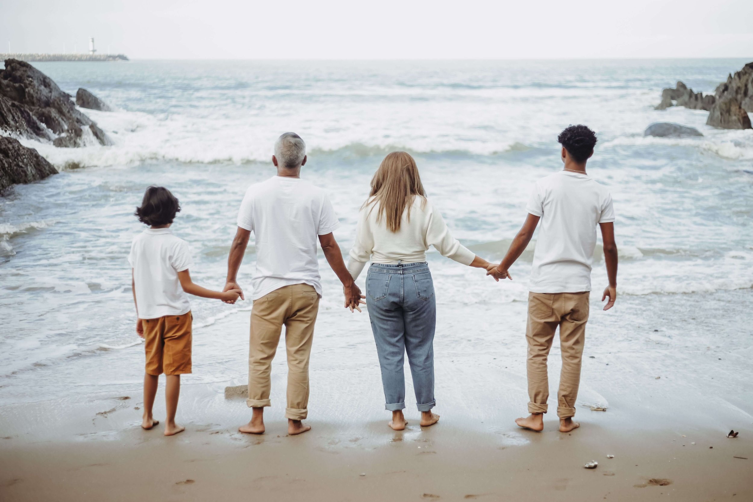 A family of 4 holding hands and looking out toward the rough waters. Representing how teen dbt therapy in Miami, FL can support the whole family. Get started with a therapist today.