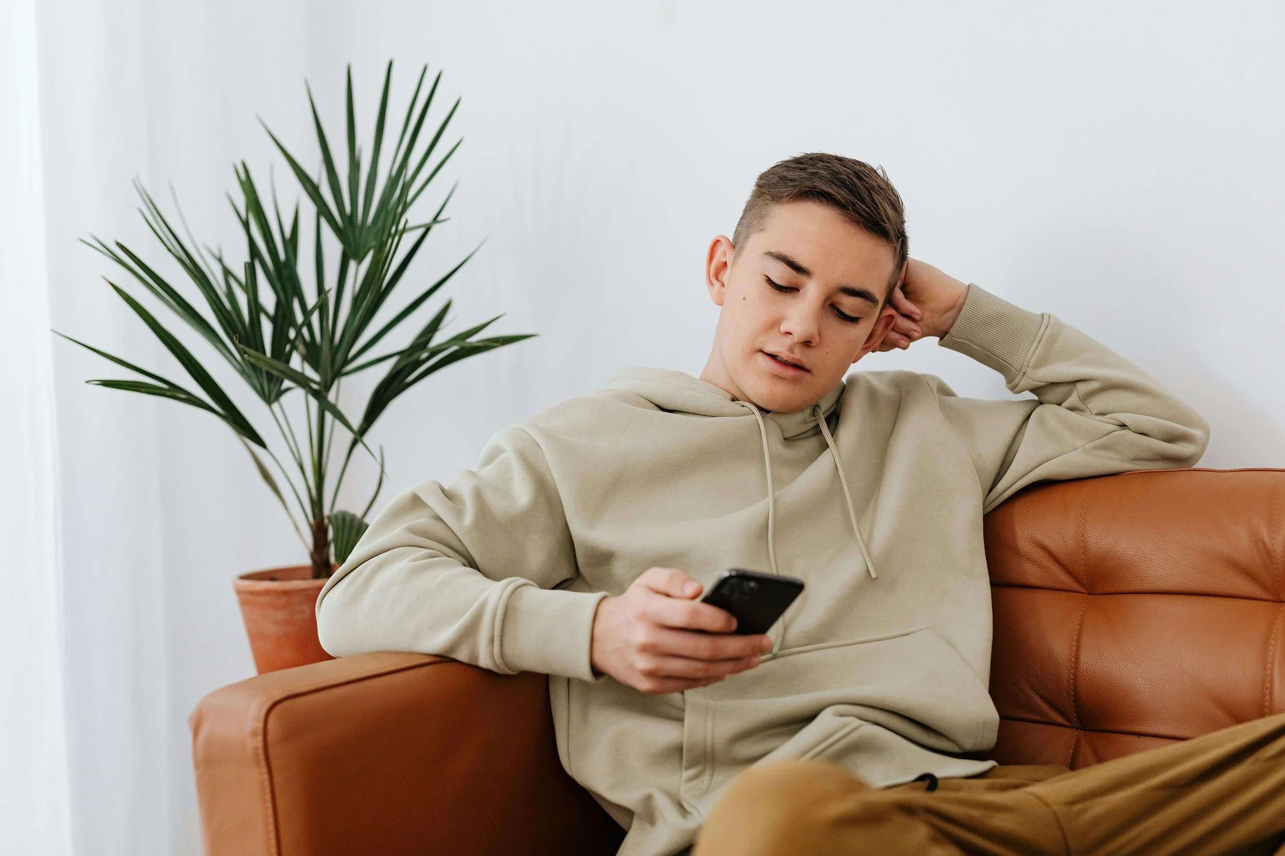 A teenage boy in a hoodie lying back on a leather couch scrolling through his phone. If your teen is spending the summer alone on the couch and glued to their phone, teen therapy in Miami, FL can help them break the cycle and find real connection.
