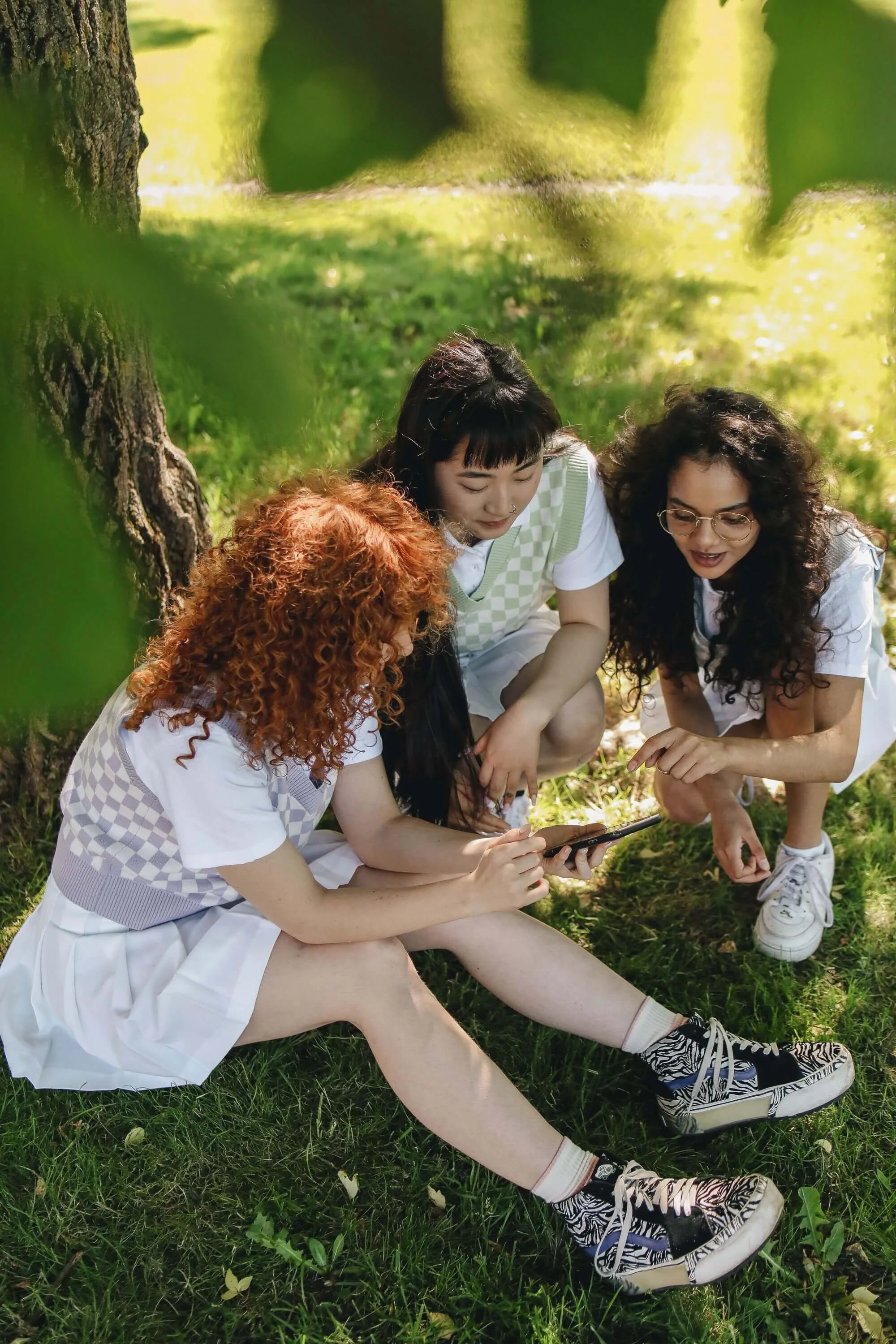 A group of diverse teen girls sitting outside near a tree in the grass looking at a phone. Teen therapy in Miami, FL is here to support your teen with ADHD management & emotional regulation. Reach out to a therapist today.