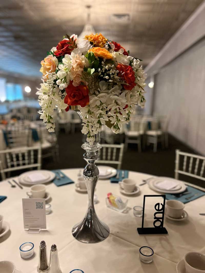 A floral centerpiece on a banquet table featuring white, red, and cream flowers with greenery in a silver vase, set for a formal event.