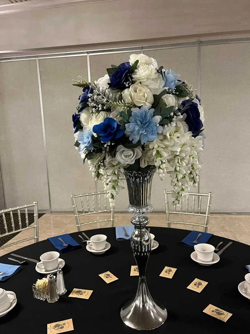 Elegant silver vase with a large bouquet of white, purple, and dark purple flowers on a black surface against a beige wall.