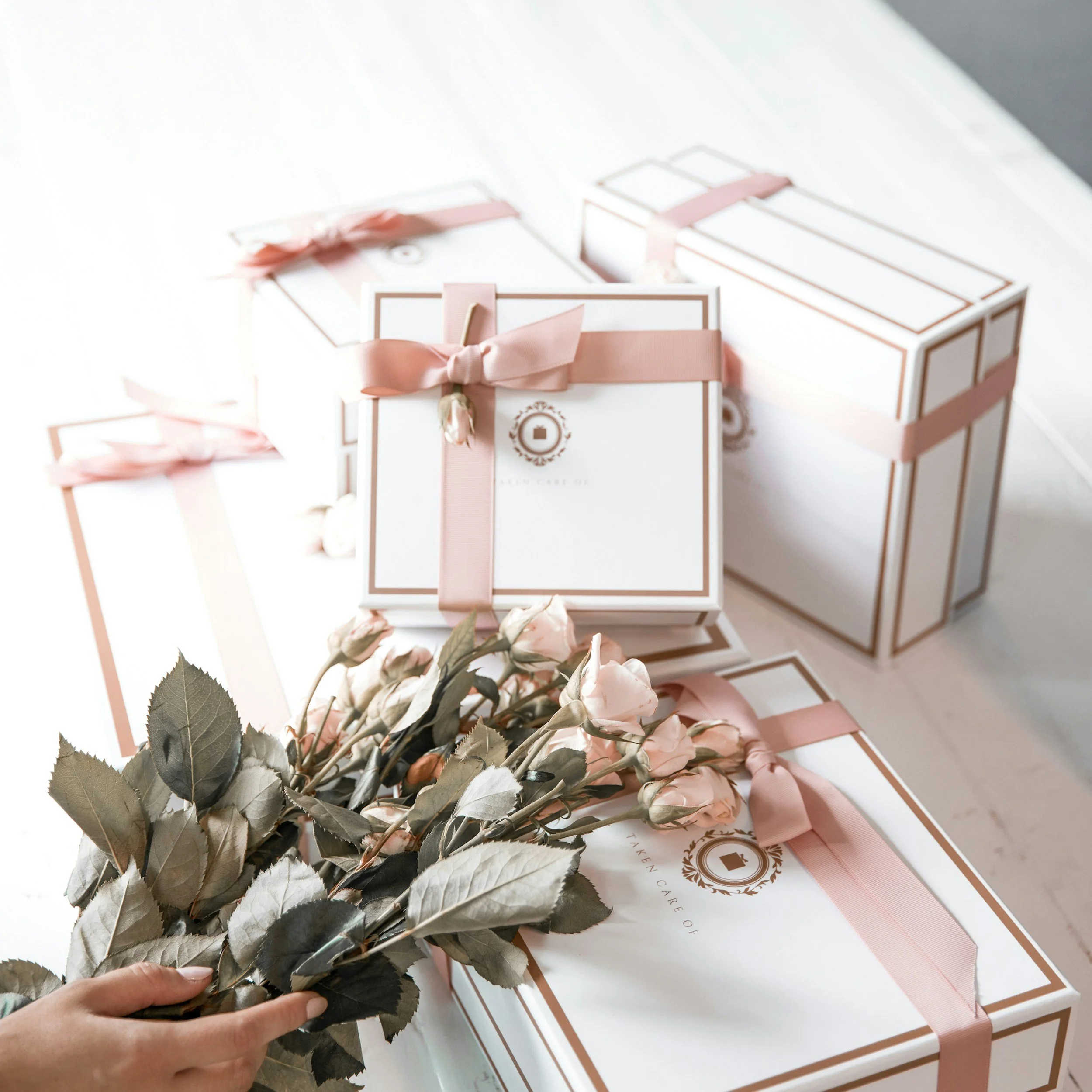 Pink and white gift boxes with ribbons, some with a logo, and a hand holding a bouquet of pink roses with gray-green leaves on a white surface.