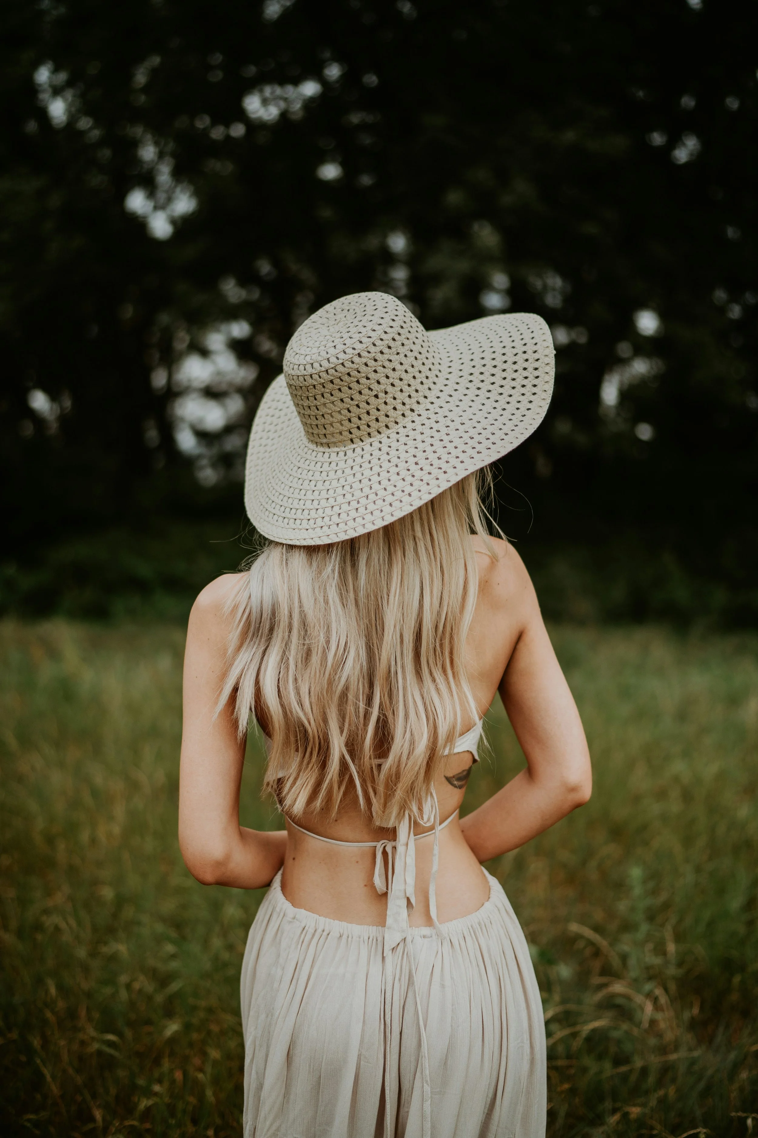 A woman with long, wavy blonde hair wearing a large, wide-brimmed straw hat and a backless top with thin straps, standing outdoors in a grassy area with trees in the background.