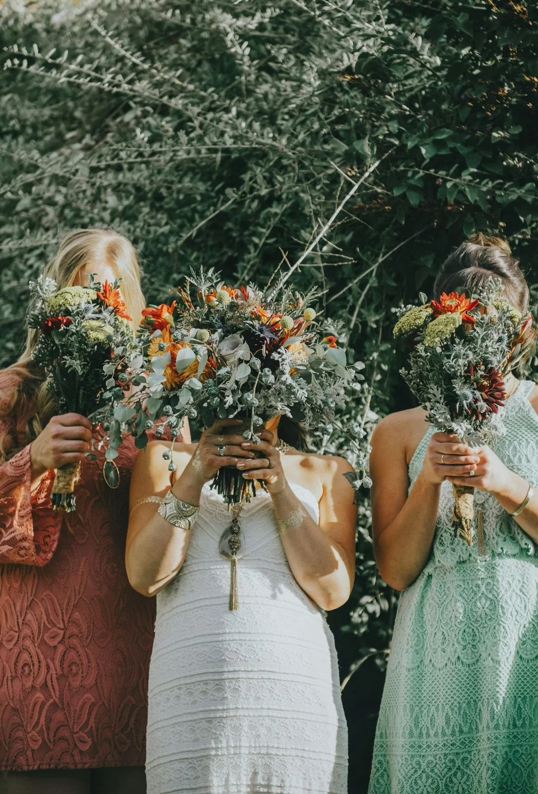 Three women holding large bouquets of colorful flowers in front of their faces during a sunny outdoor event.