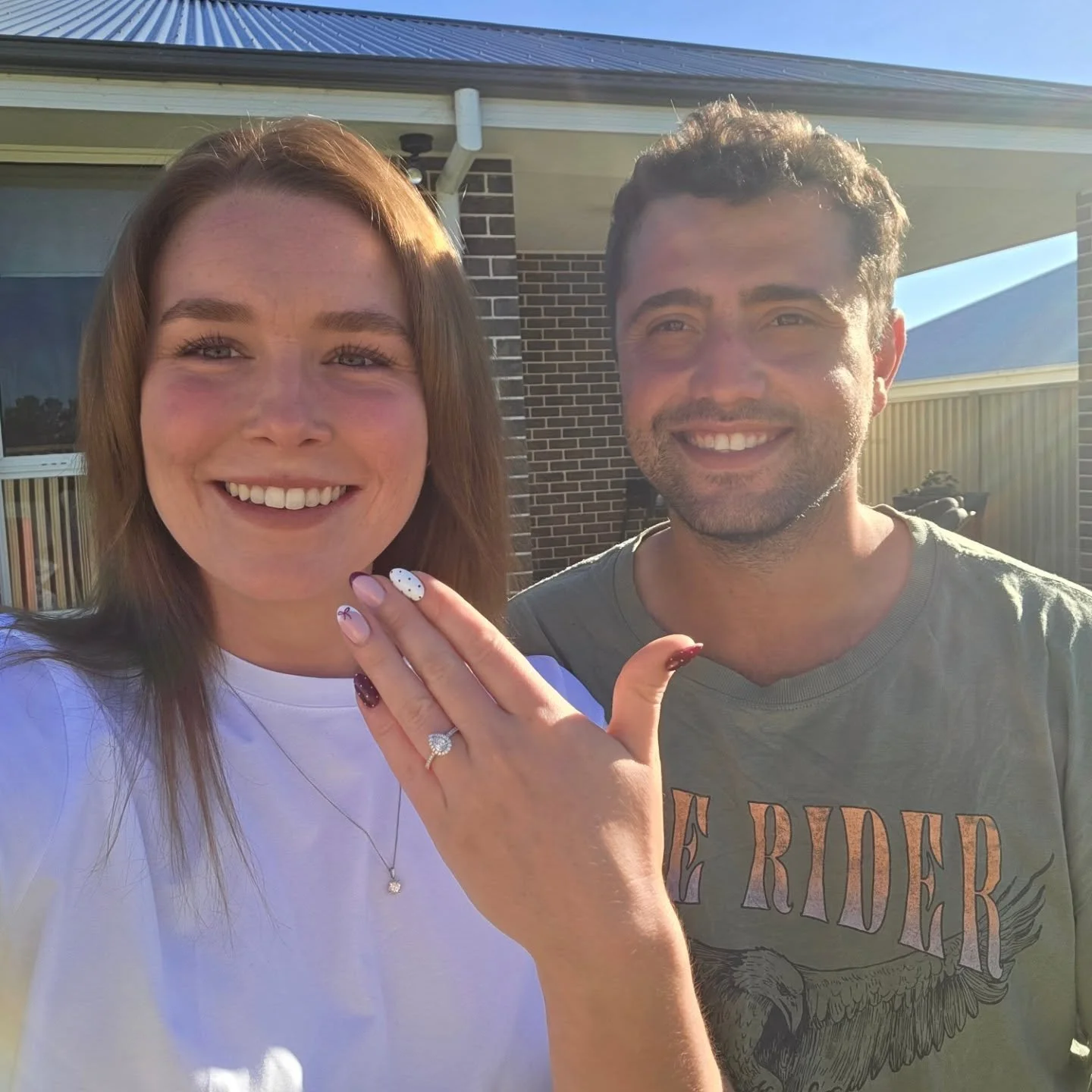 Liv, Clean Livin's Director,  is smiling and showing her engagement ring and Jesse, Liv's husband, standing outdoors in bright sunlight, in front of a house with brick and siding, during daytime.