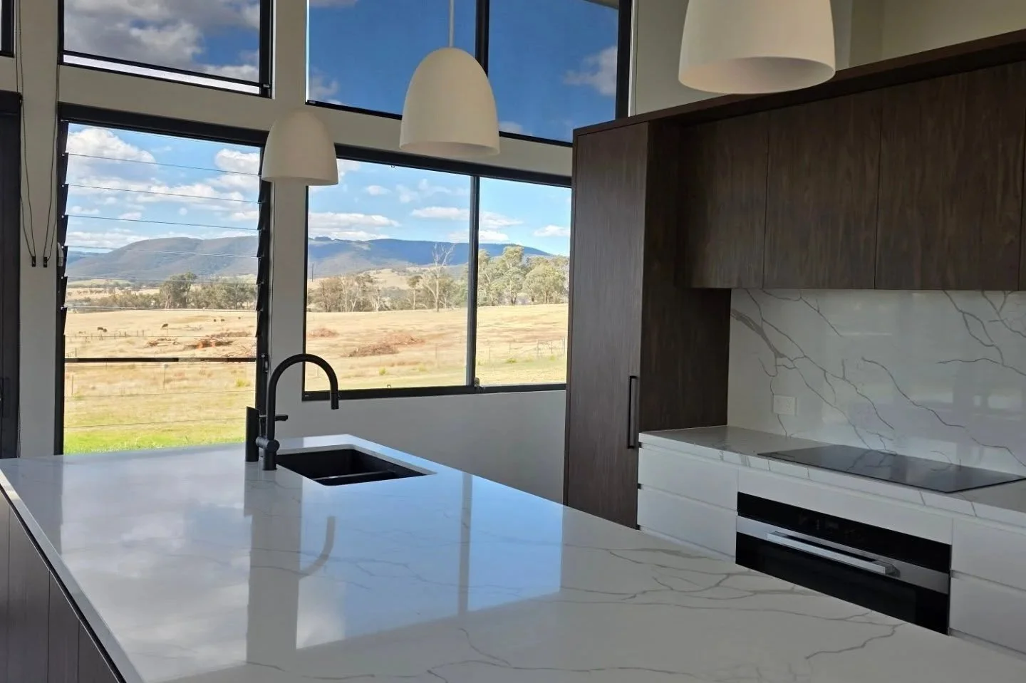 Modern kitchen with white marble countertops, dark wood cabinets, black sink, and large windows showing a scenic landscape with mountains and open fields.